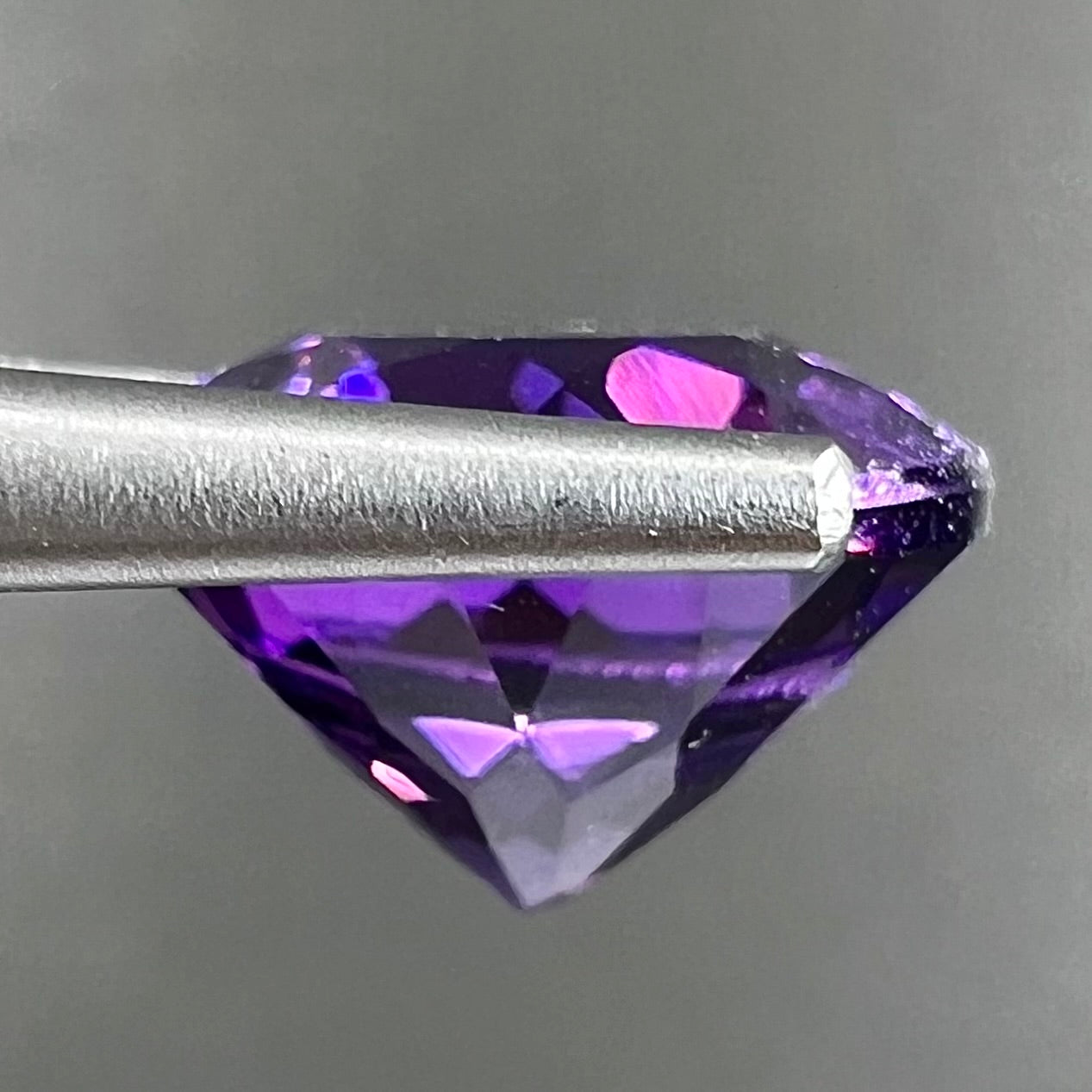 A loose, faceted round brilliant cut amethyst gemstone. The stone is purple with blue flashes.