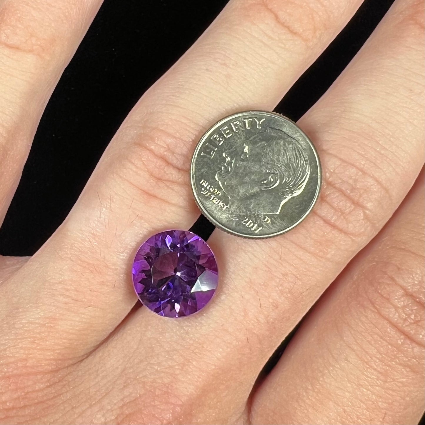 A loose, faceted round brilliant cut amethyst gemstone. The stone is purple with blue flashes.