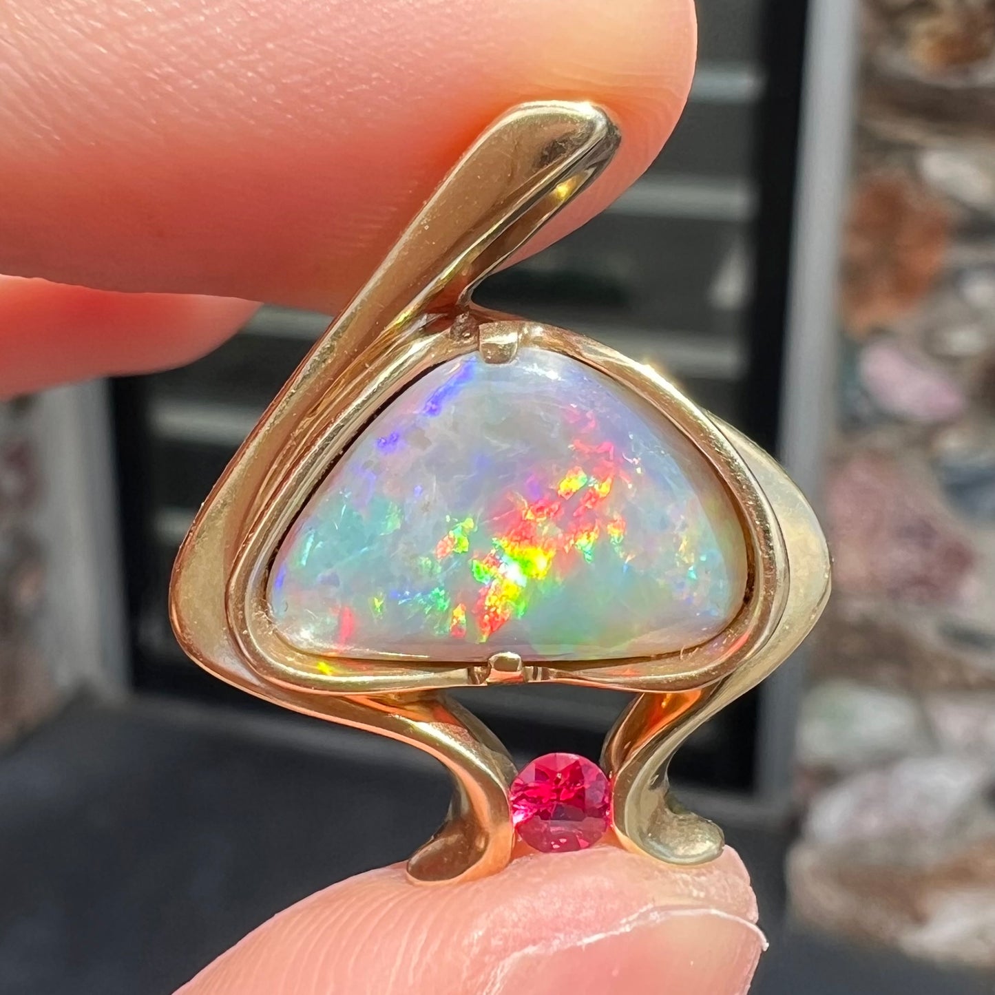 A ladies' yellow gold pendant bezel set with a natural black opal and pink sapphire accent. The opal shines red, green, blue, and purple.
