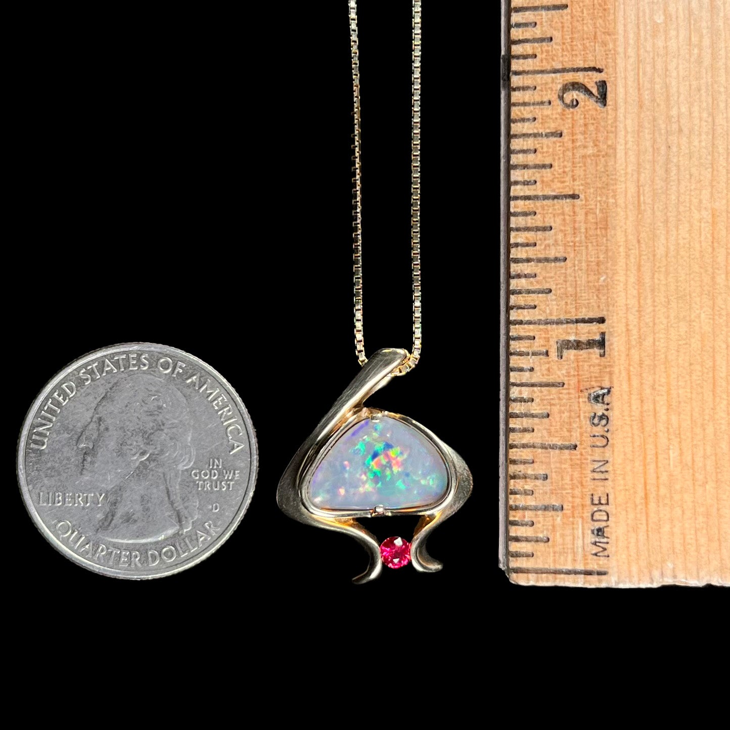 A ladies' yellow gold pendant bezel set with a natural black opal and pink sapphire accent. The opal shines red, green, blue, and purple.