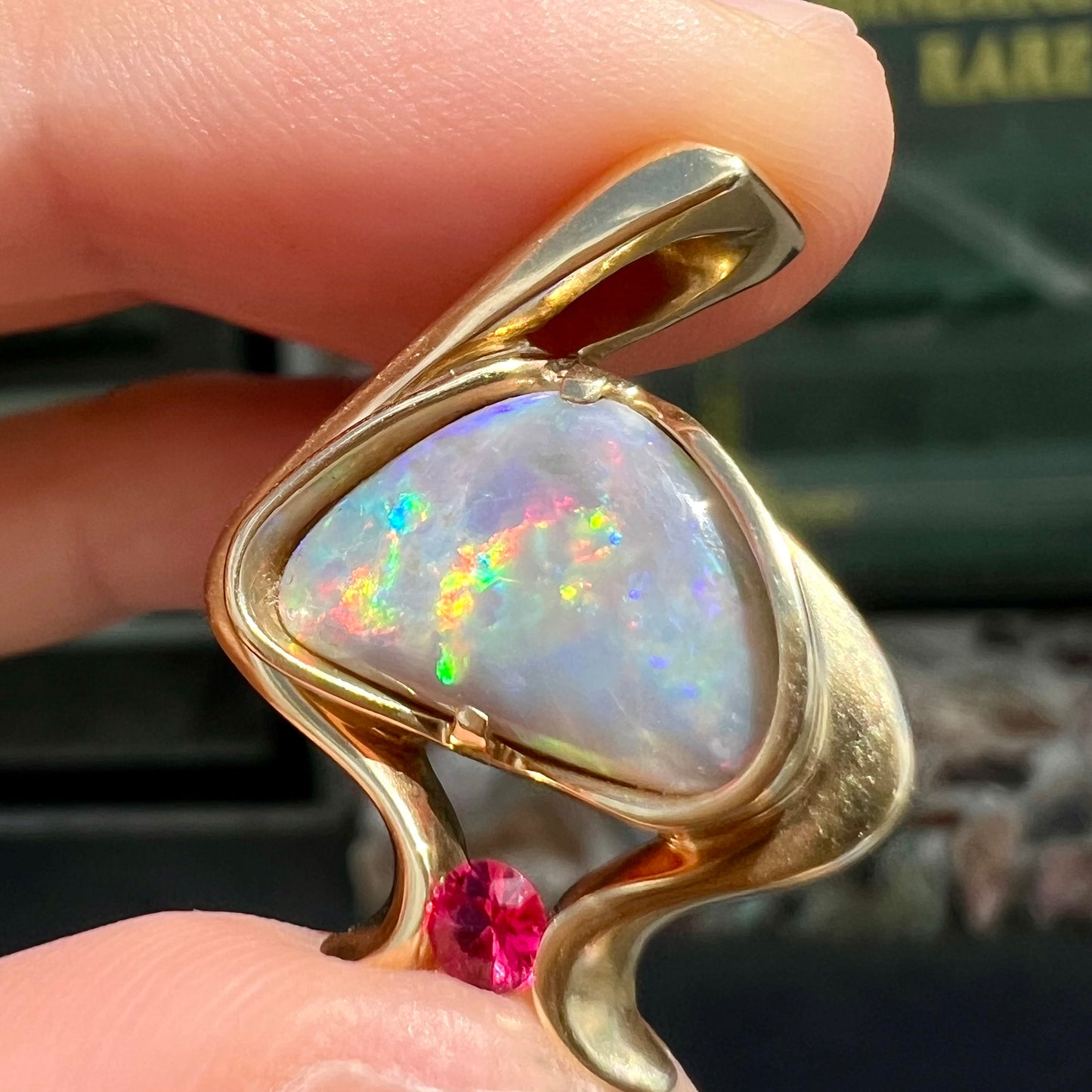 A ladies' yellow gold pendant bezel set with a natural black opal and pink sapphire accent. The opal shines red, green, blue, and purple.