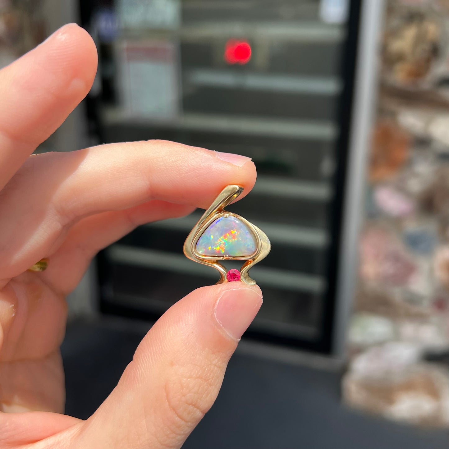 A ladies' yellow gold pendant bezel set with a natural black opal and pink sapphire accent. The opal shines red, green, blue, and purple.
