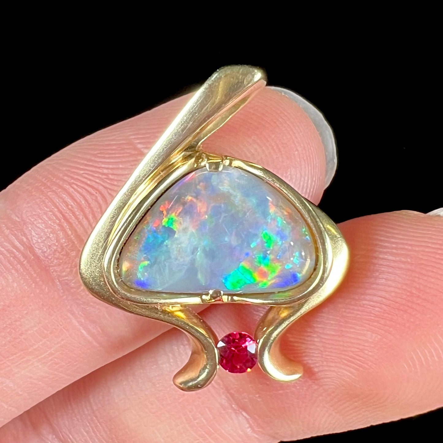 A ladies' yellow gold pendant bezel set with a natural black opal and pink sapphire accent. The opal shines red, green, blue, and purple.