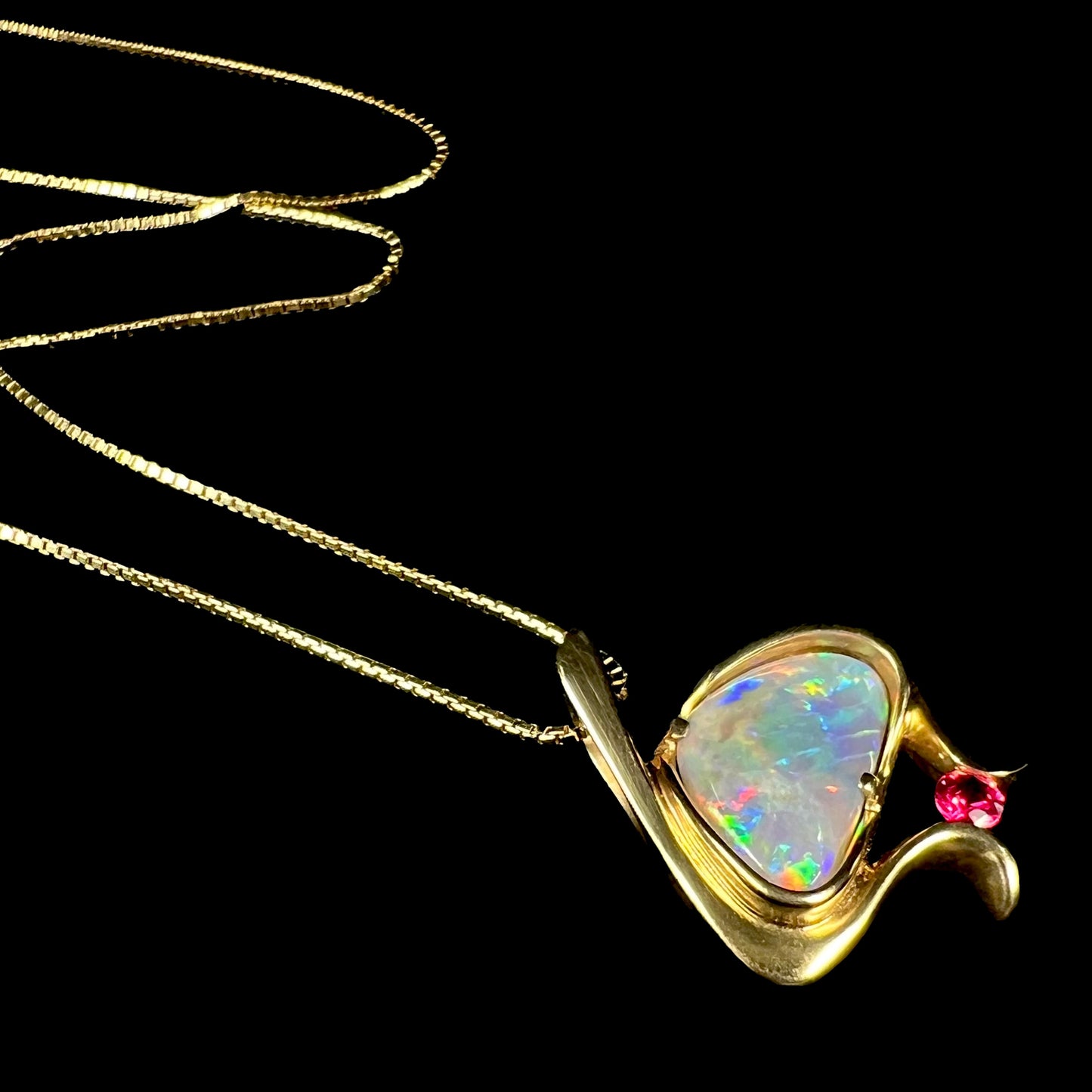 A ladies' yellow gold pendant bezel set with a natural black opal and pink sapphire accent. The opal shines red, green, blue, and purple.
