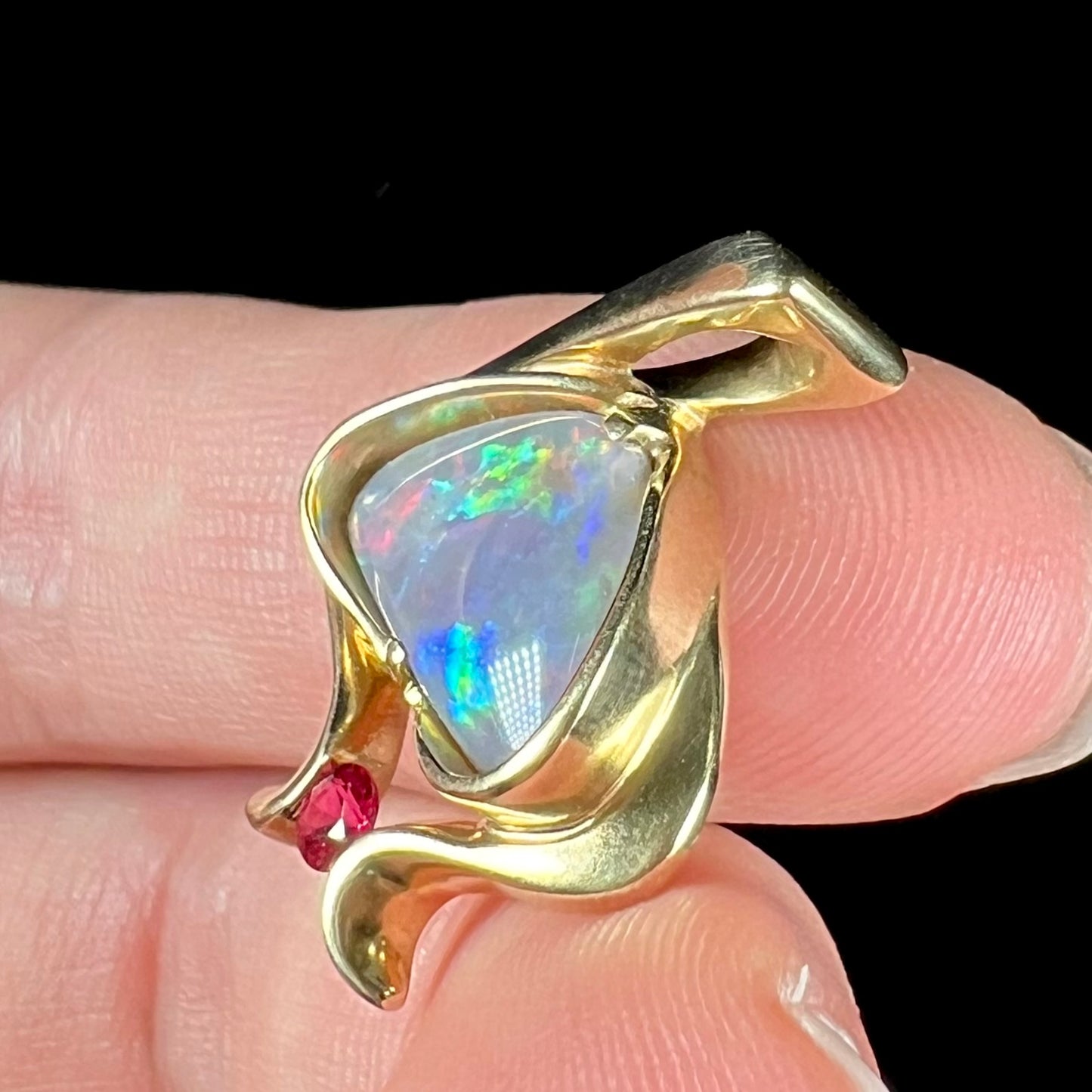 A ladies' yellow gold pendant bezel set with a natural black opal and pink sapphire accent. The opal shines red, green, blue, and purple.