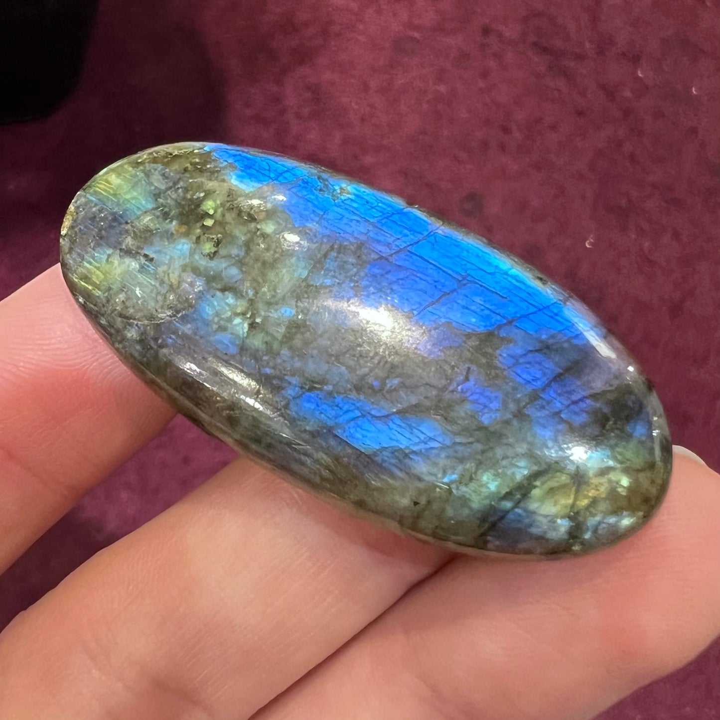 A loose, oval cabochon cut labradorite stone. The stone is green with bright blue and yellow flashes.