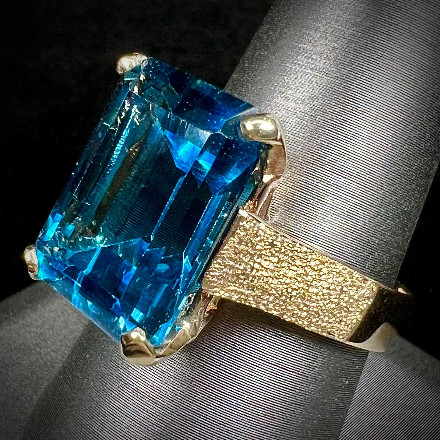 A ladies' yellow gold London blue topaz solitaire ring. The ring is textured, and the topaz is emerald cut.