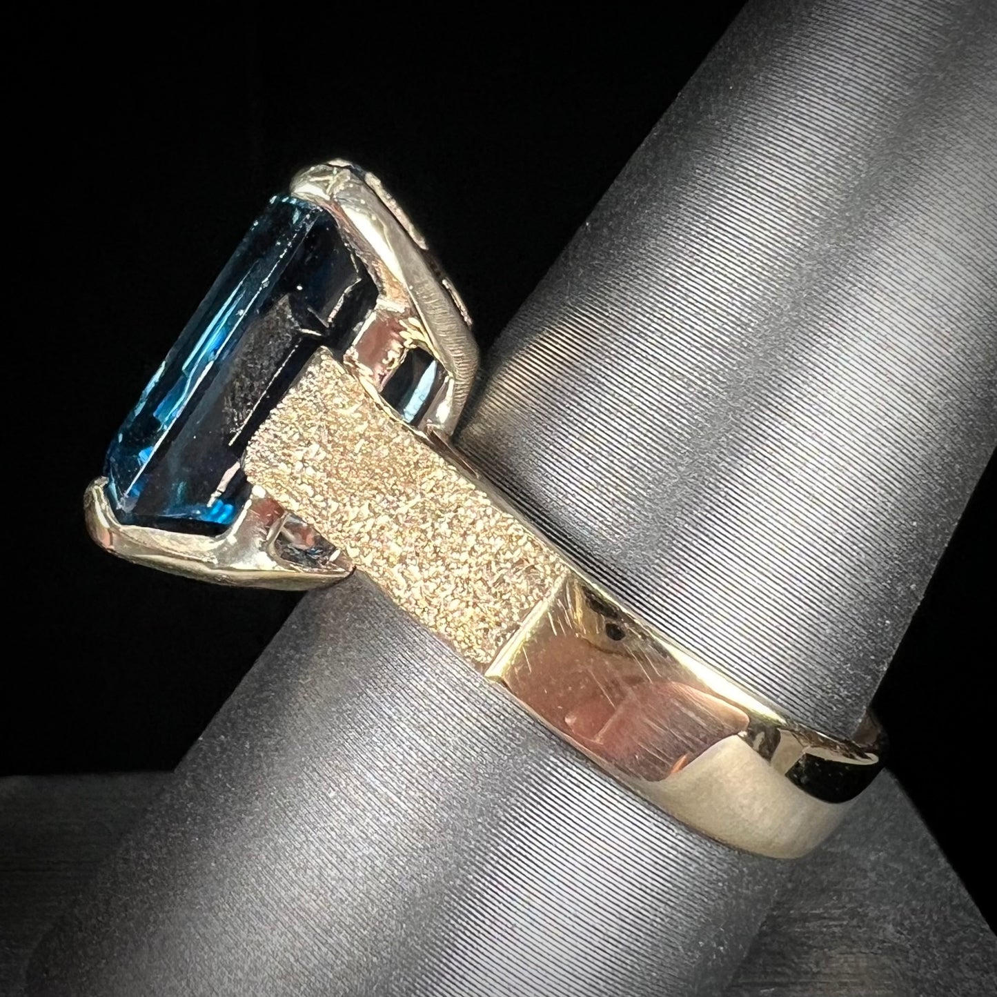 A ladies' yellow gold London blue topaz solitaire ring. The ring is textured, and the topaz is emerald cut.