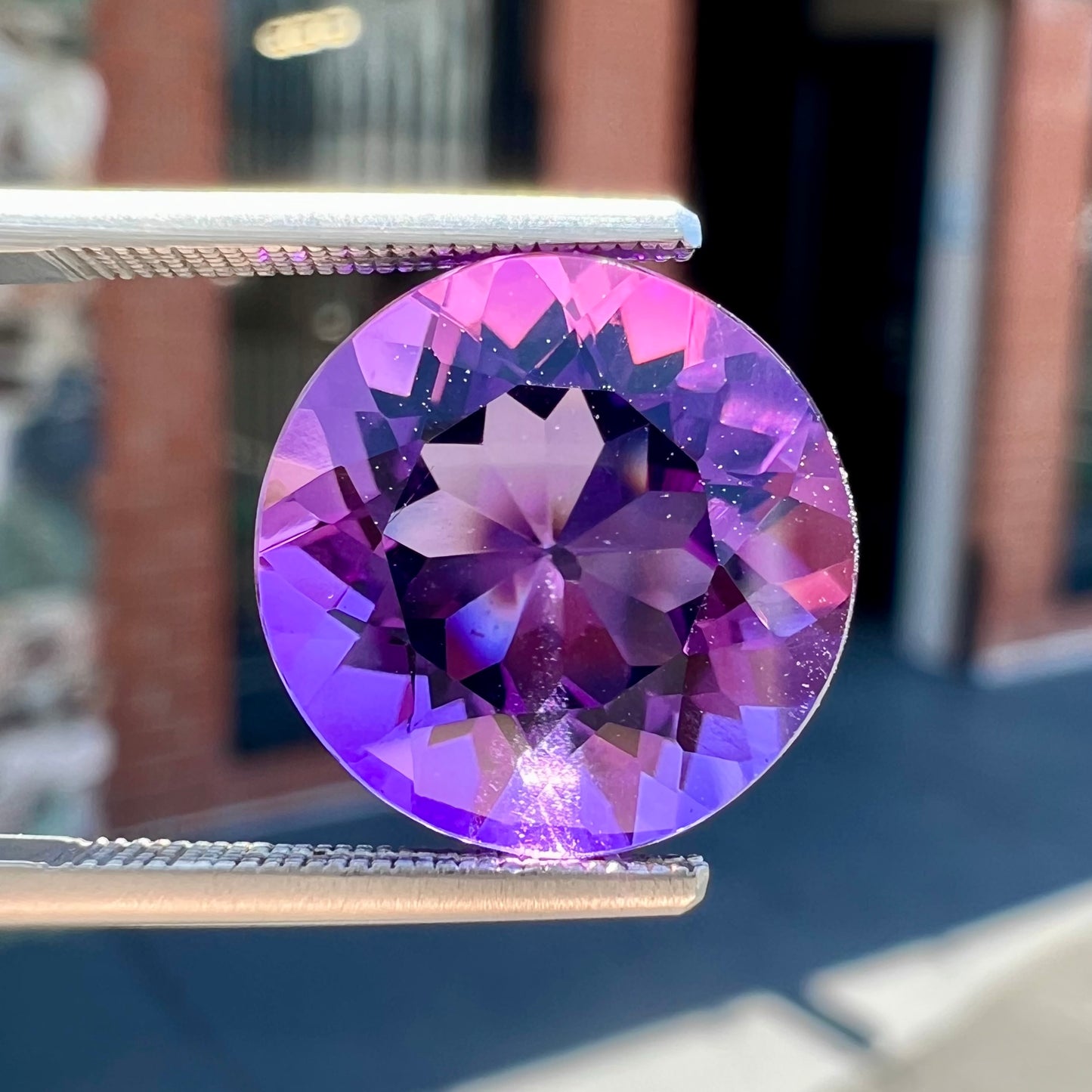 A loose, faceted round brilliant cut amethyst gemstone. The stone is purple with blue flashes.