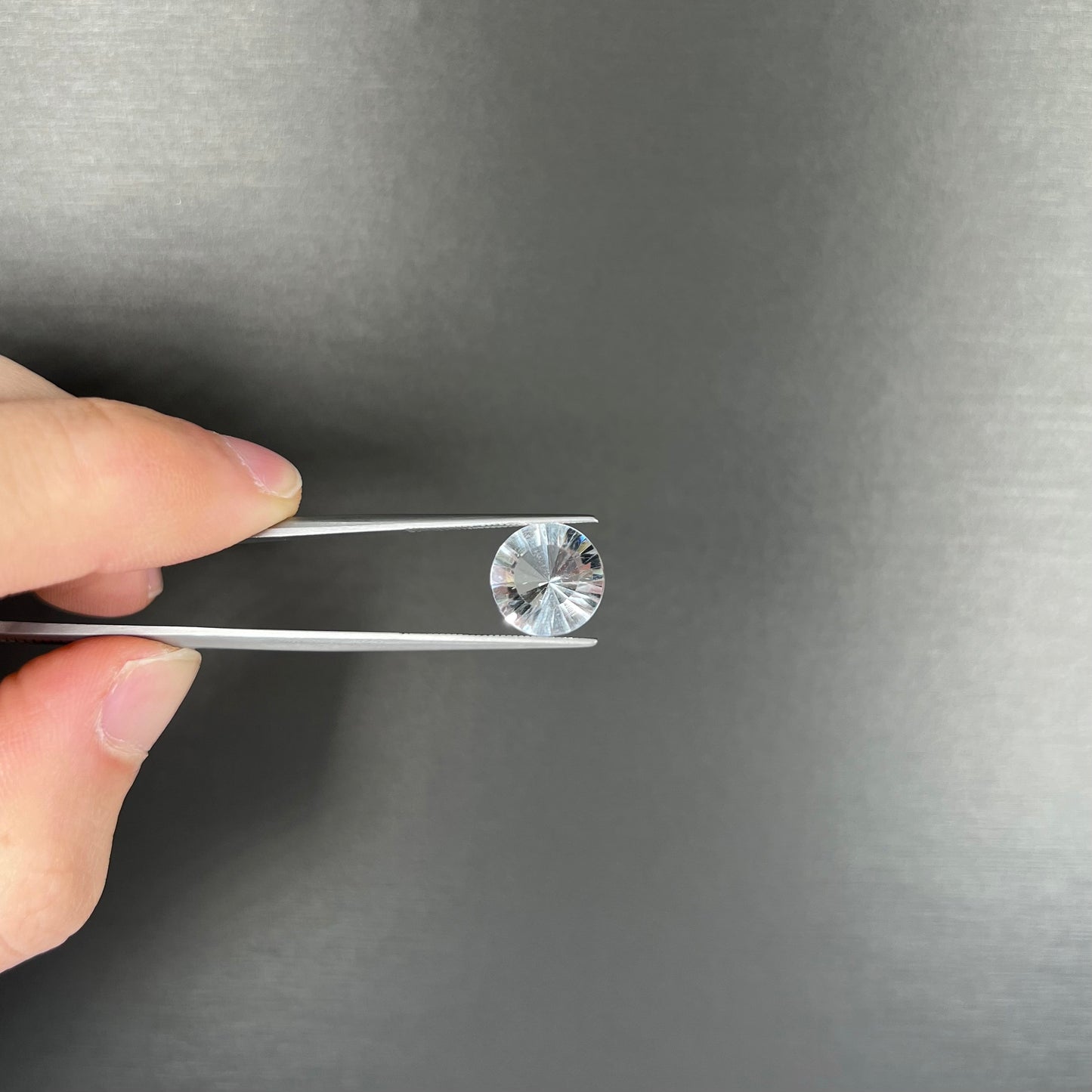 A loose, round cut petalite gemstone. The faceted stone is colorless with a black surface inclusion.