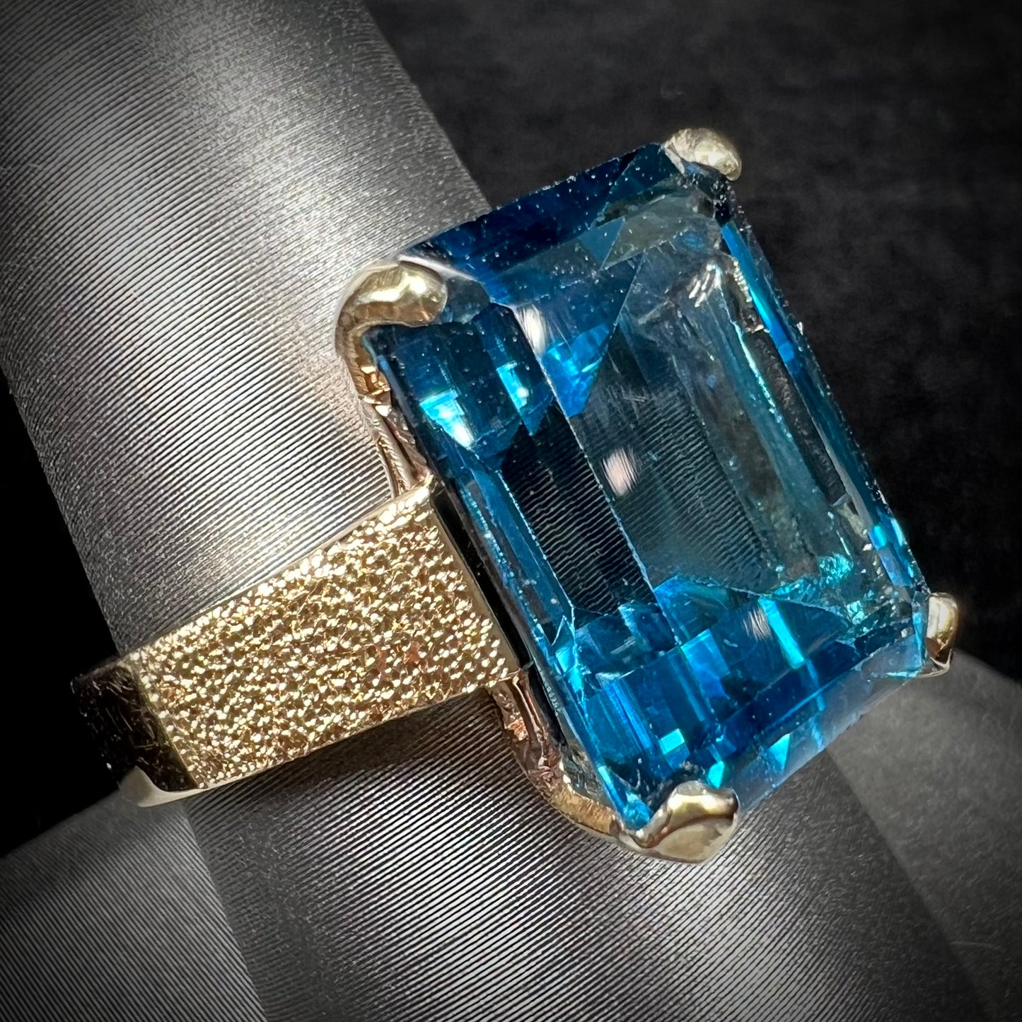 A ladies' yellow gold London blue topaz solitaire ring. The ring is textured, and the topaz is emerald cut.
