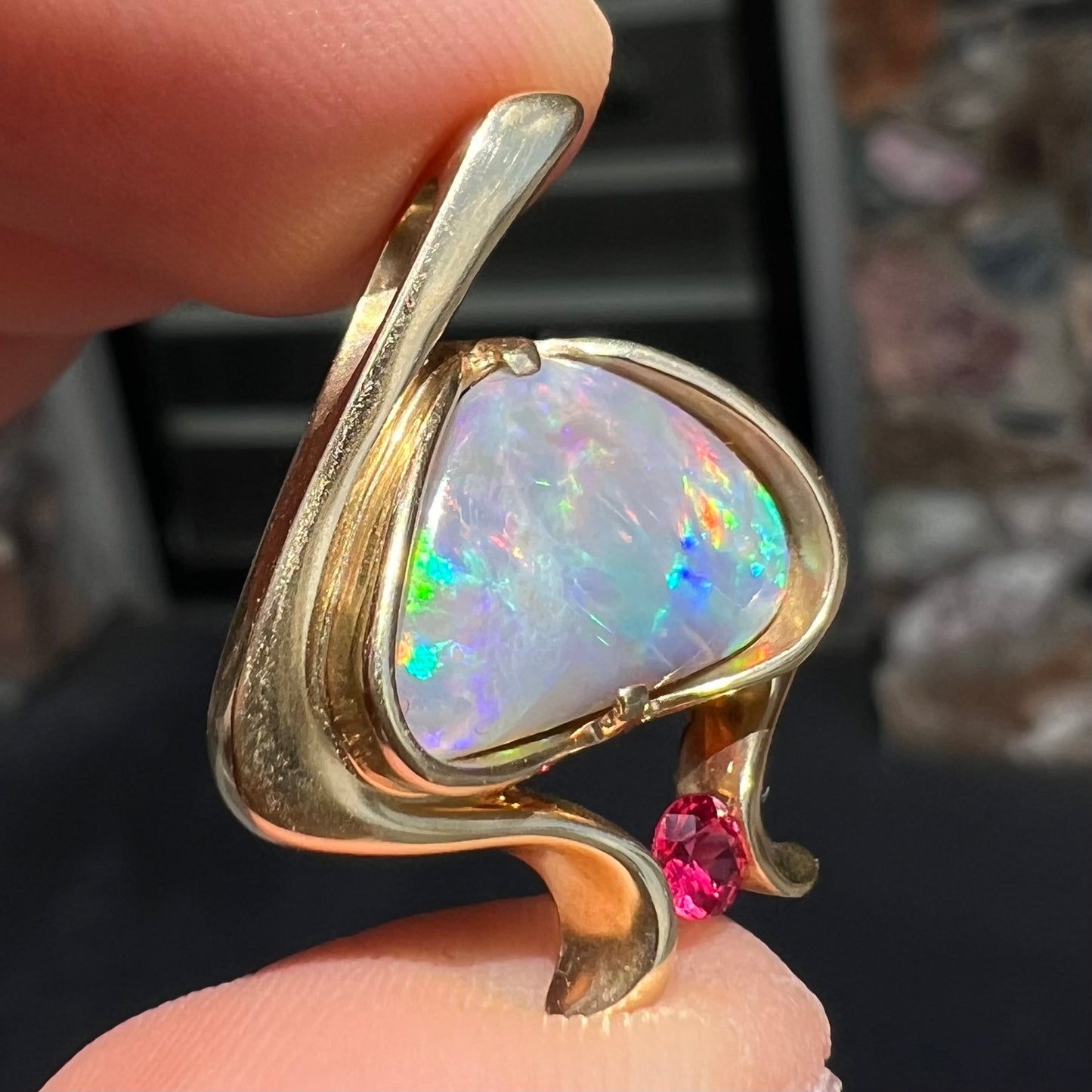 A ladies' yellow gold pendant bezel set with a natural black opal and pink sapphire accent. The opal shines red, green, blue, and purple.