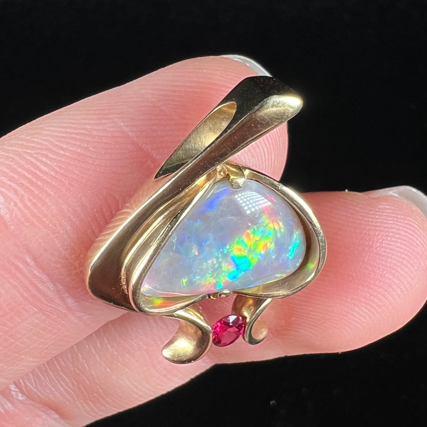 A ladies' yellow gold pendant bezel set with a natural black opal and pink sapphire accent. The opal shines red, green, blue, and purple.