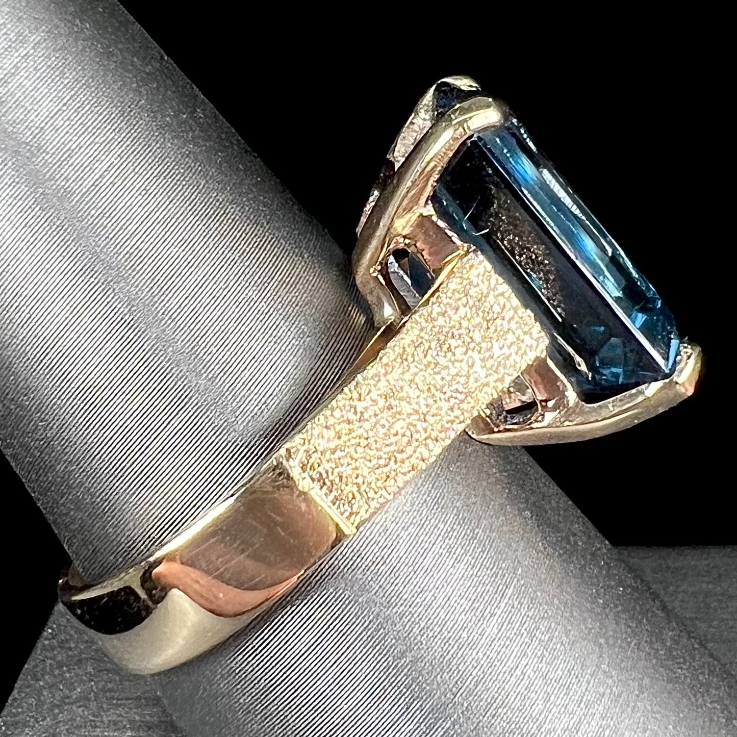 A ladies' yellow gold London blue topaz solitaire ring. The ring is textured, and the topaz is emerald cut.