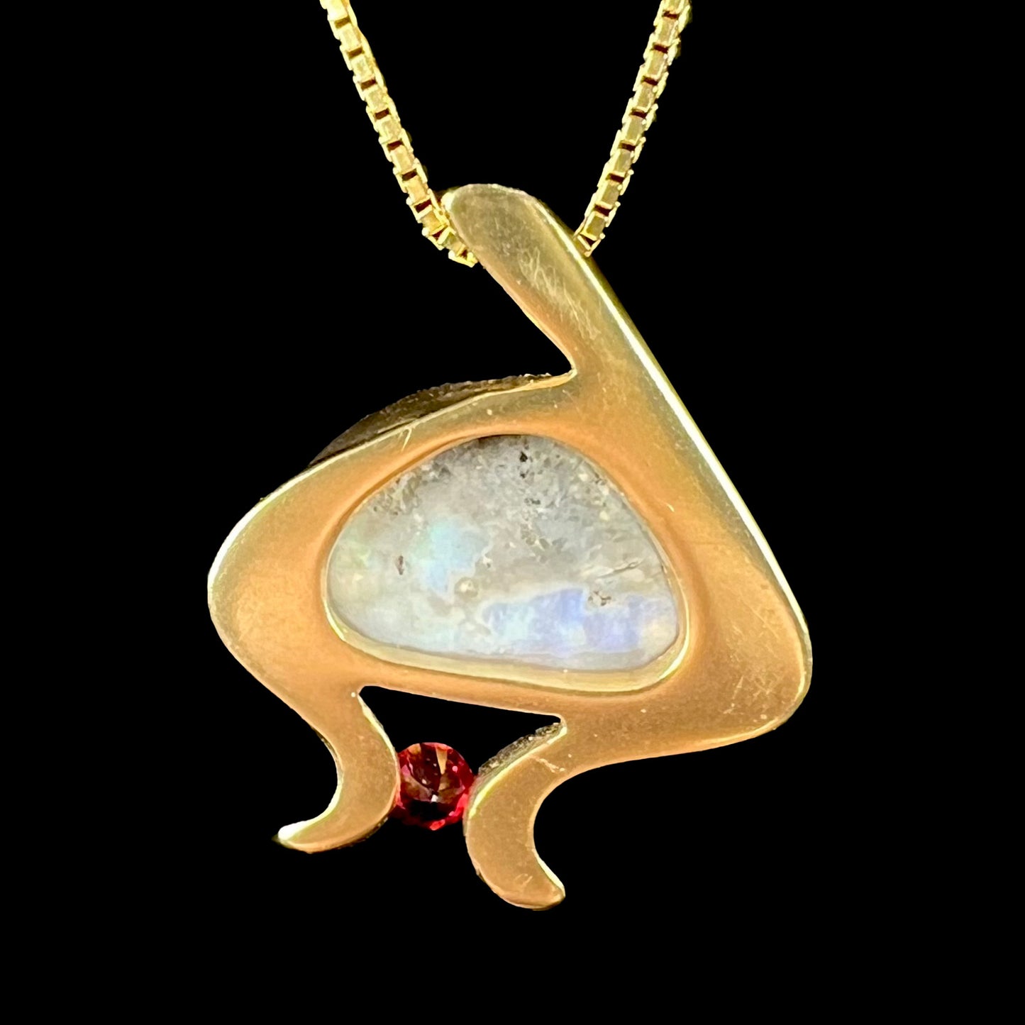 A ladies' yellow gold pendant bezel set with a natural black opal and pink sapphire accent. The opal shines red, green, blue, and purple.