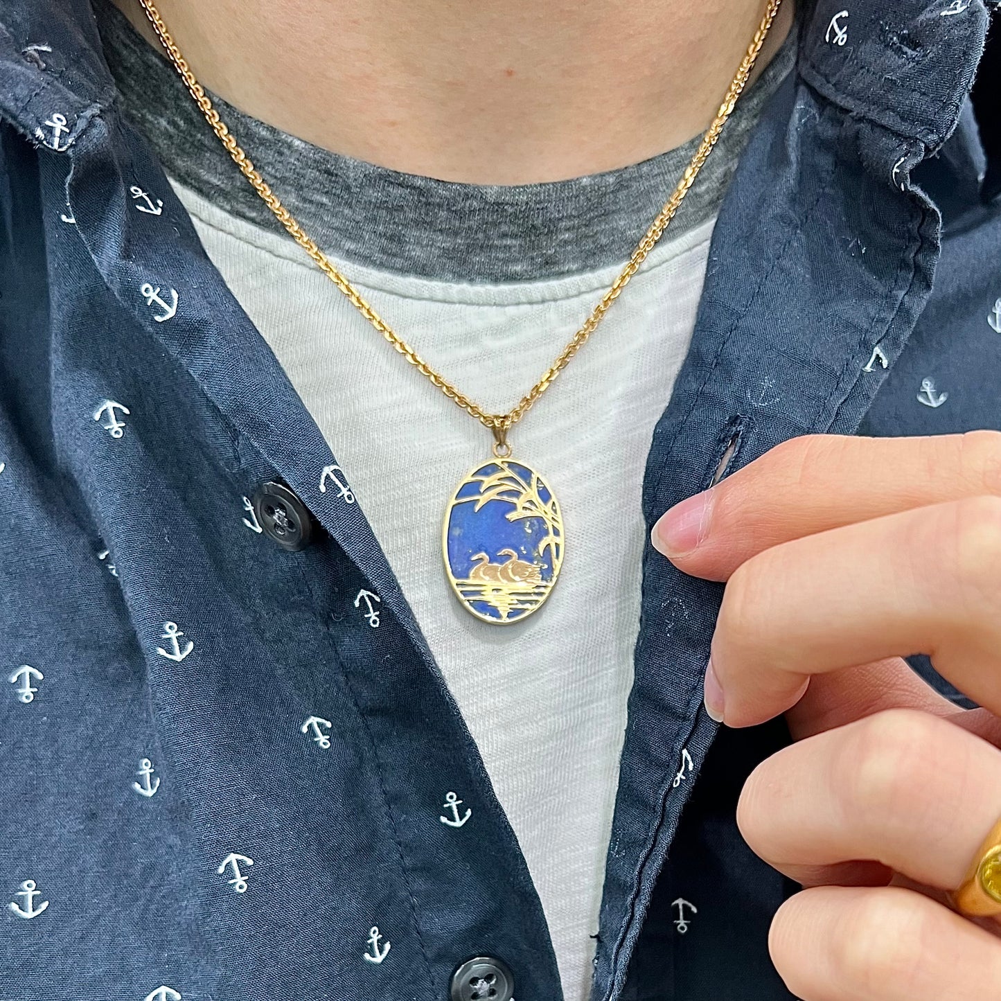 An oval cut lapis lazuli pendant set in a yellow gold frame featuring the design of two swans swimming in a pond.