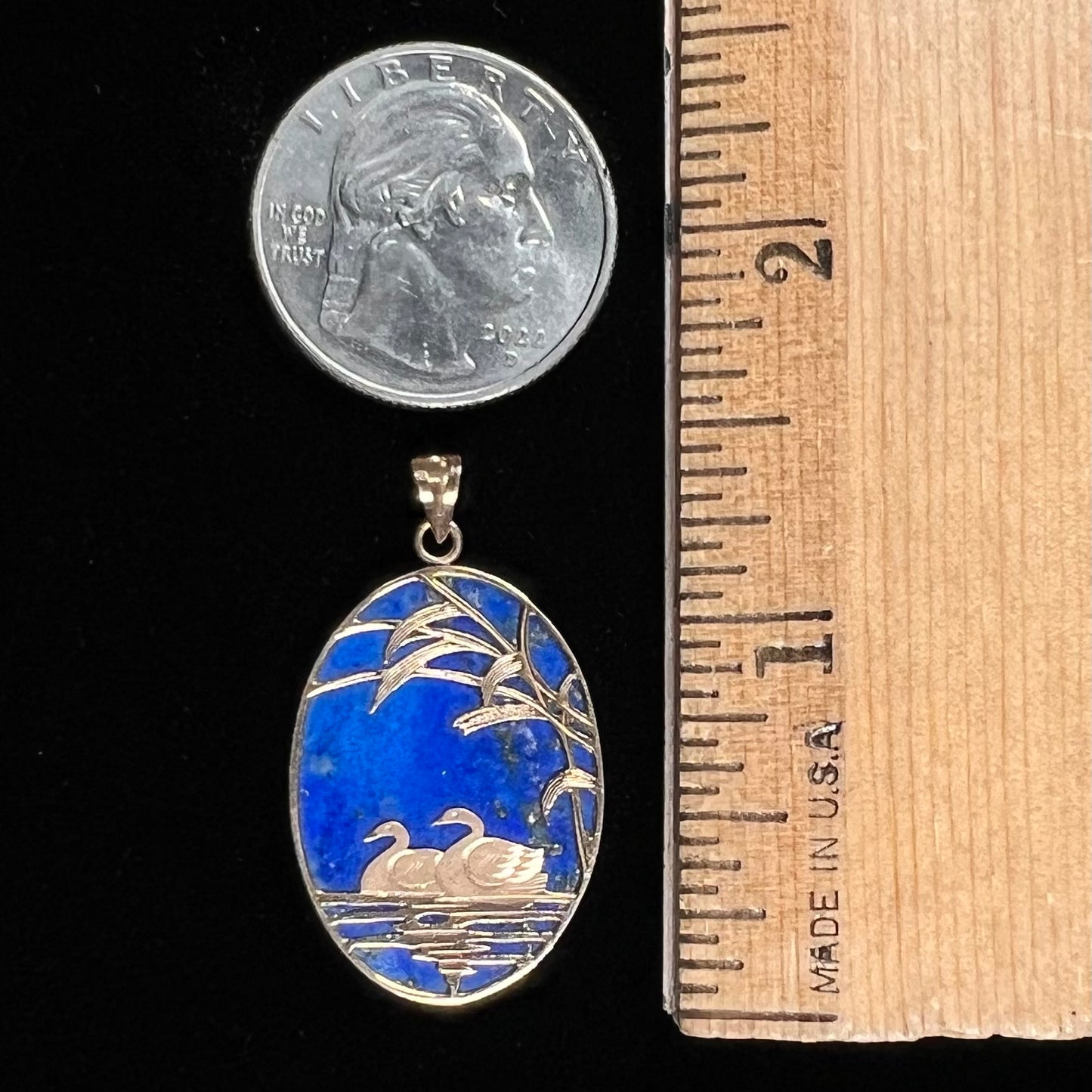 An oval cut lapis lazuli pendant set in a yellow gold frame featuring the design of two swans swimming in a pond.