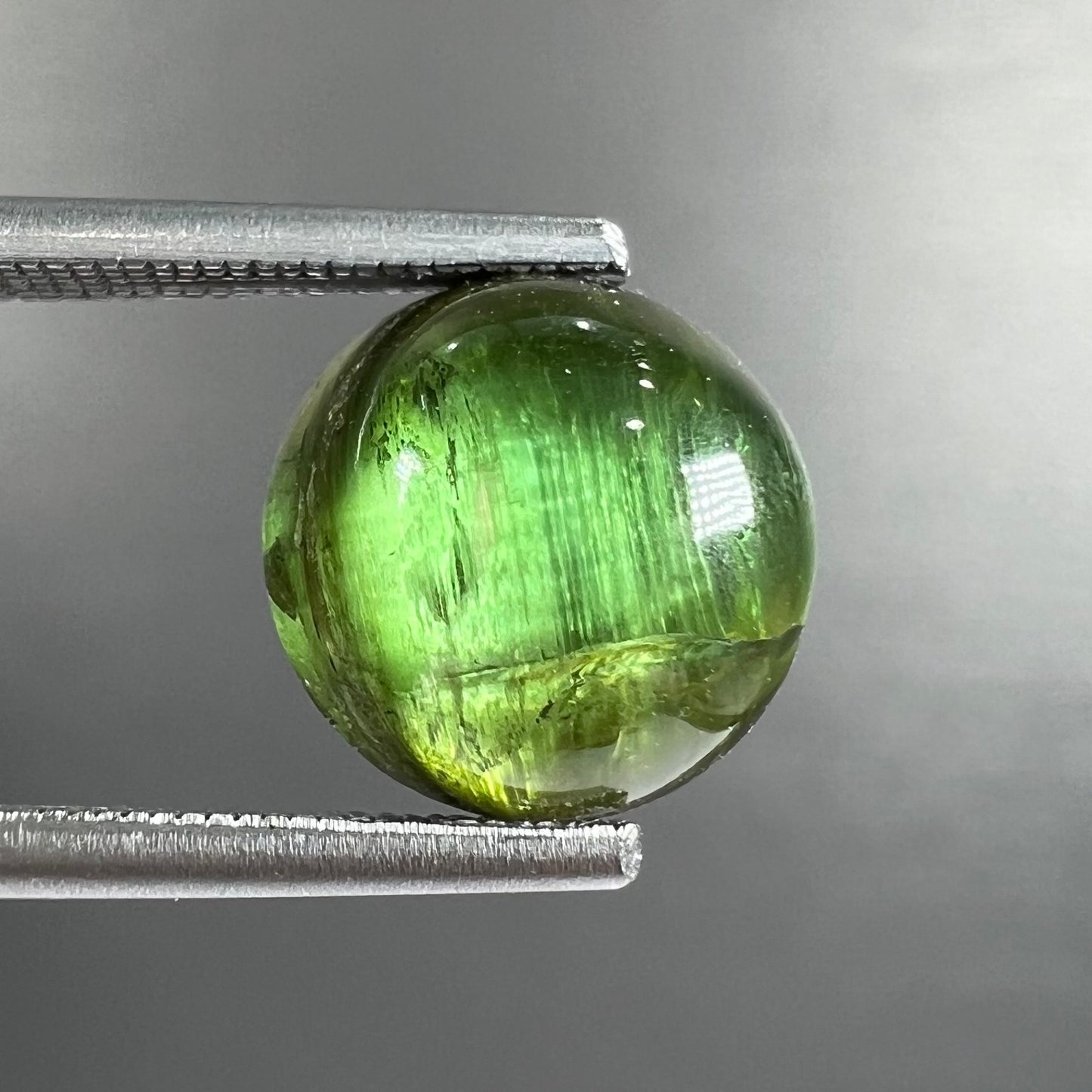 A loose, round cabochon cut green tourmaline stone that displays a cat's eye phenomenon.