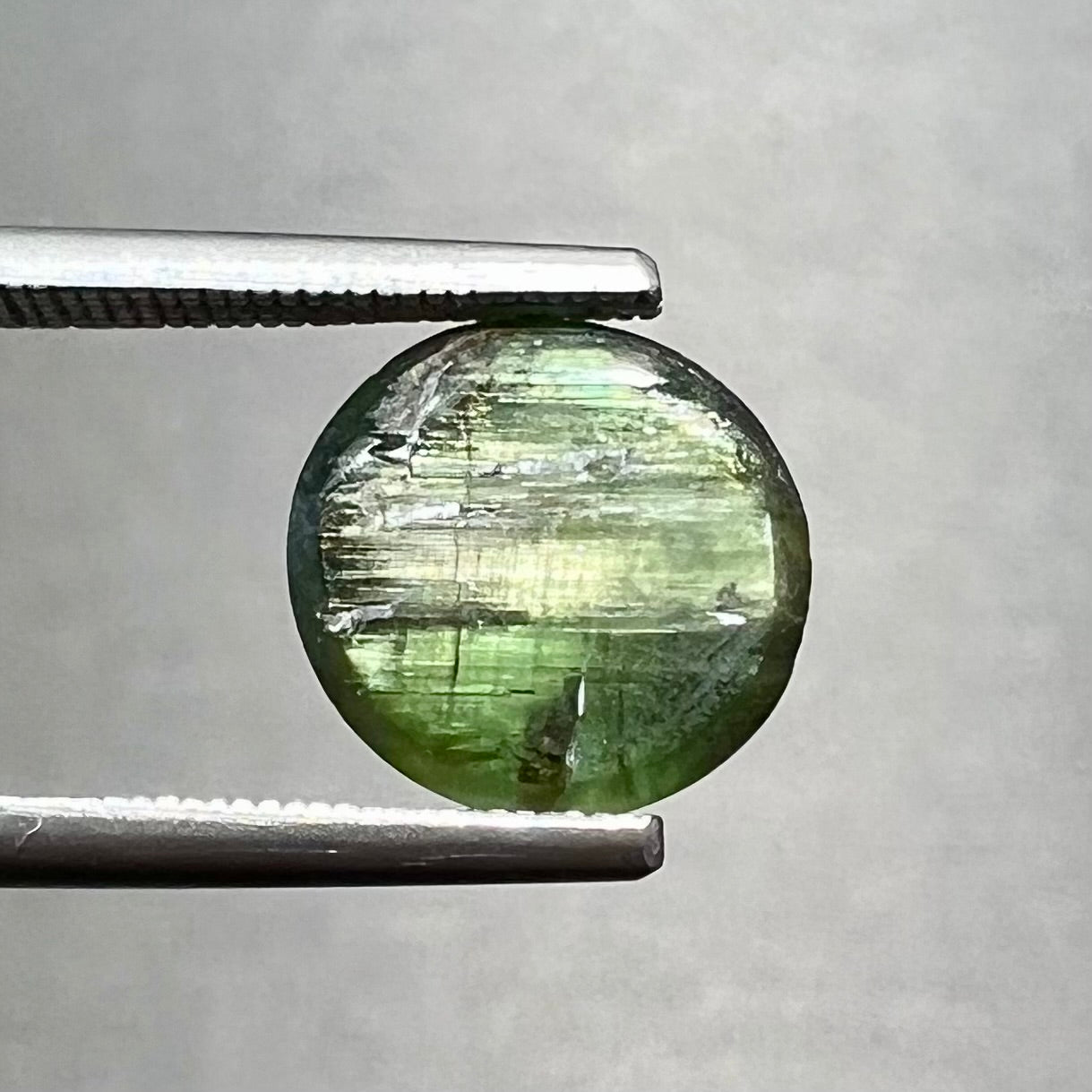 A loose, round cabochon cut green tourmaline stone that displays a cat's eye phenomenon.
