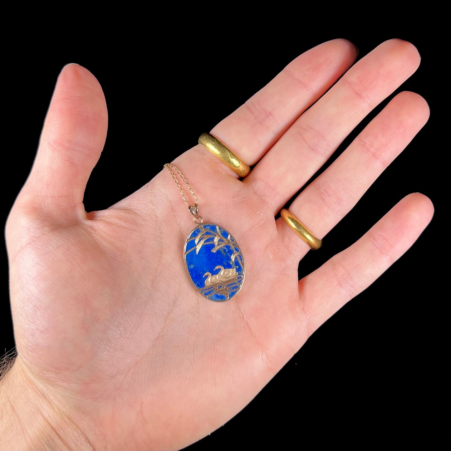 An oval cut lapis lazuli pendant set in a yellow gold frame featuring the design of two swans swimming in a pond.