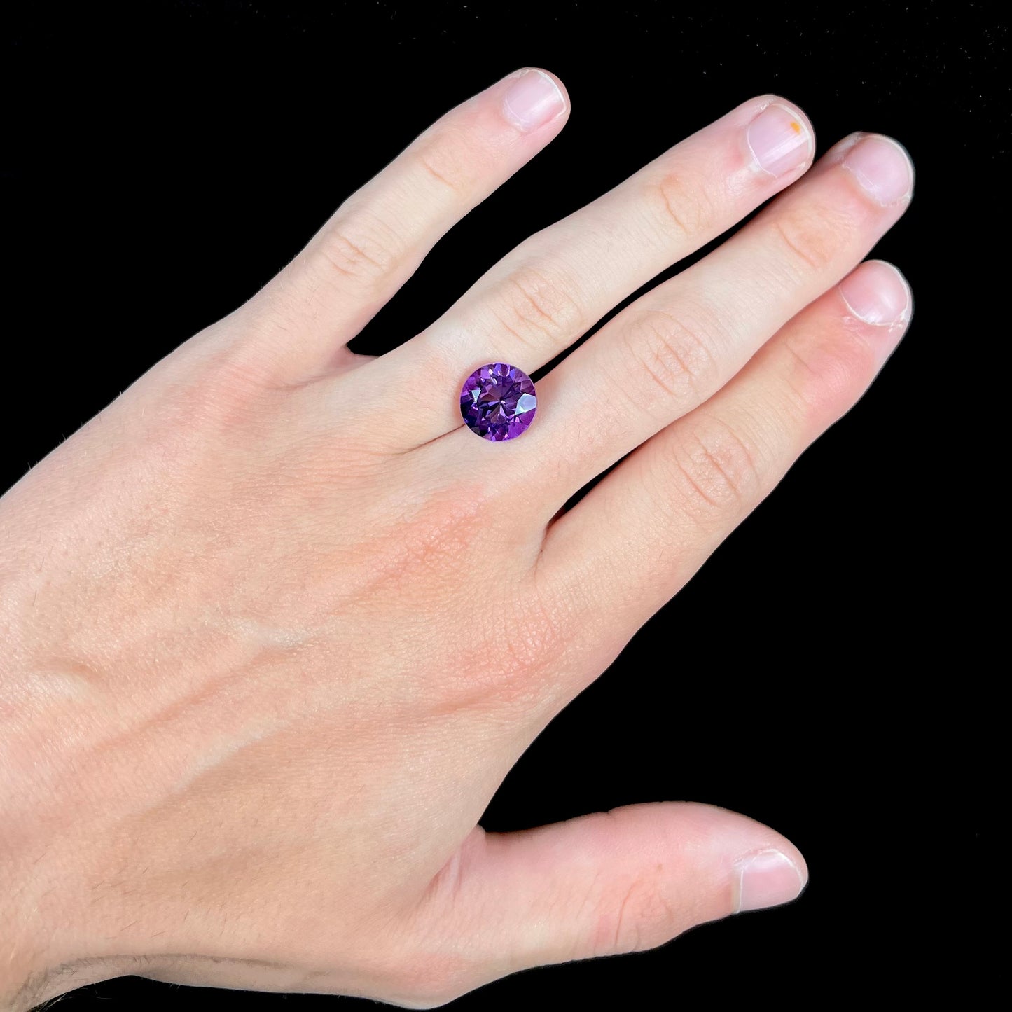 A loose, faceted round brilliant cut amethyst gemstone. The stone is purple with blue flashes.