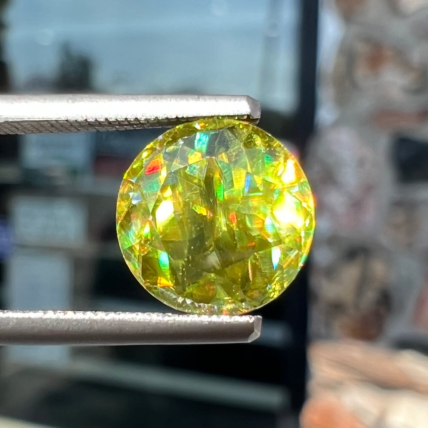 A loose, round brilliant cut sphene gemstone. The stone is green with orange, green, blue, and yellow flashes.