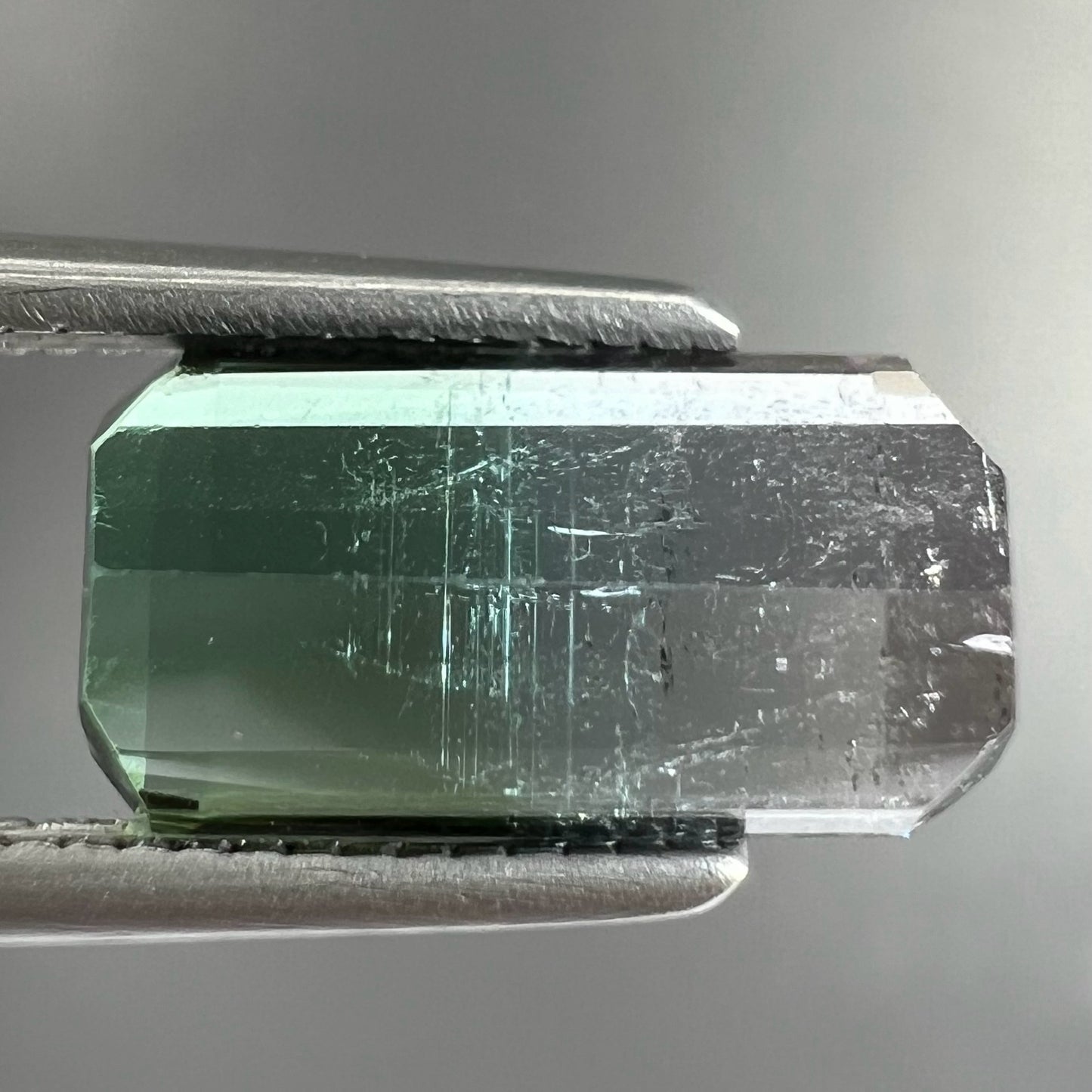 A loose, emerald cut tricolor tourmaline gemstone. The stone transitions from green to blue to white colors.