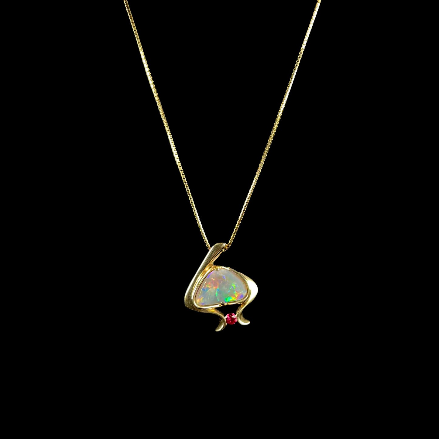A ladies' yellow gold pendant bezel set with a natural black opal and pink sapphire accent. The opal shines red, green, blue, and purple.