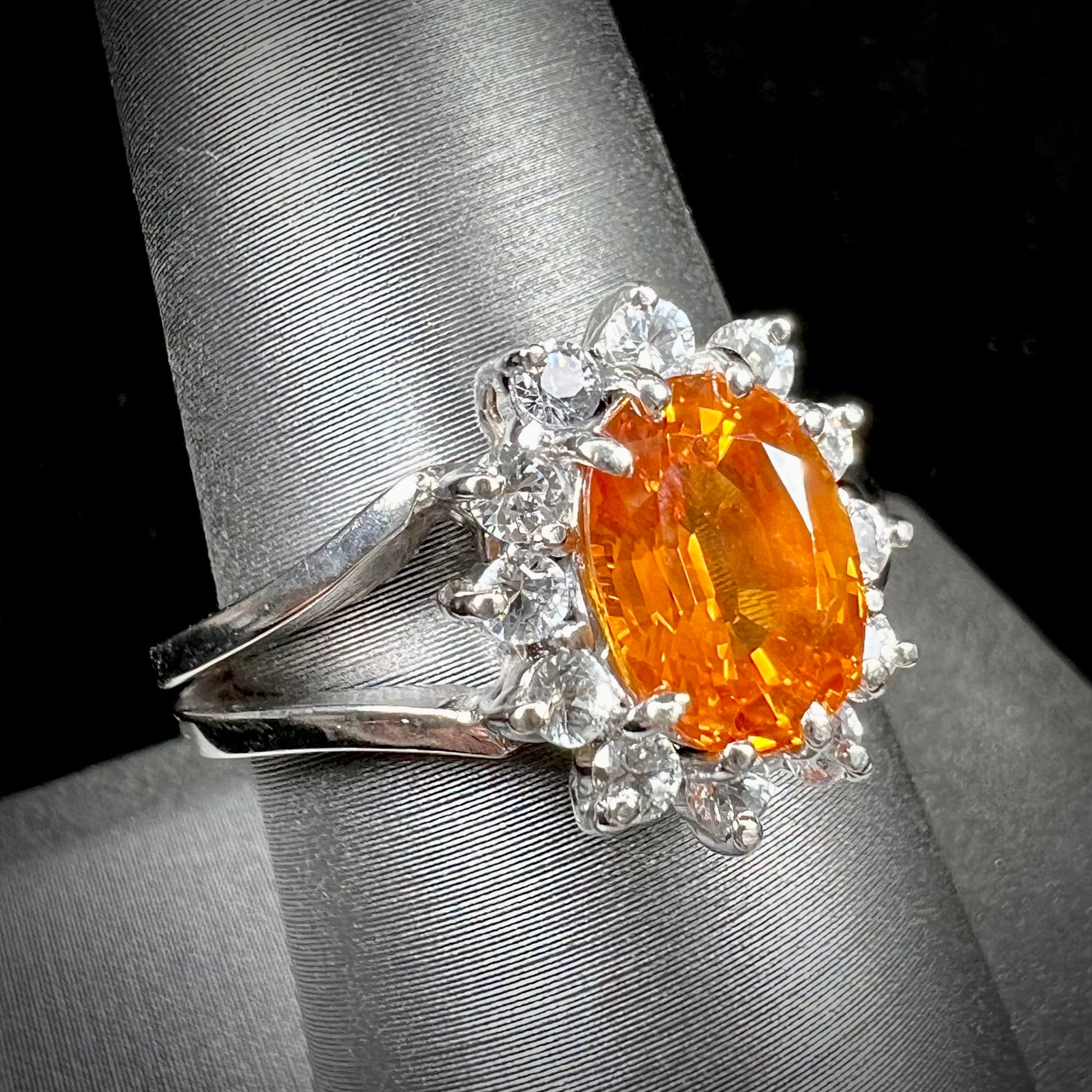 A white gold split shank ring set with an oval cut orange sapphire and a halo of white sapphires.