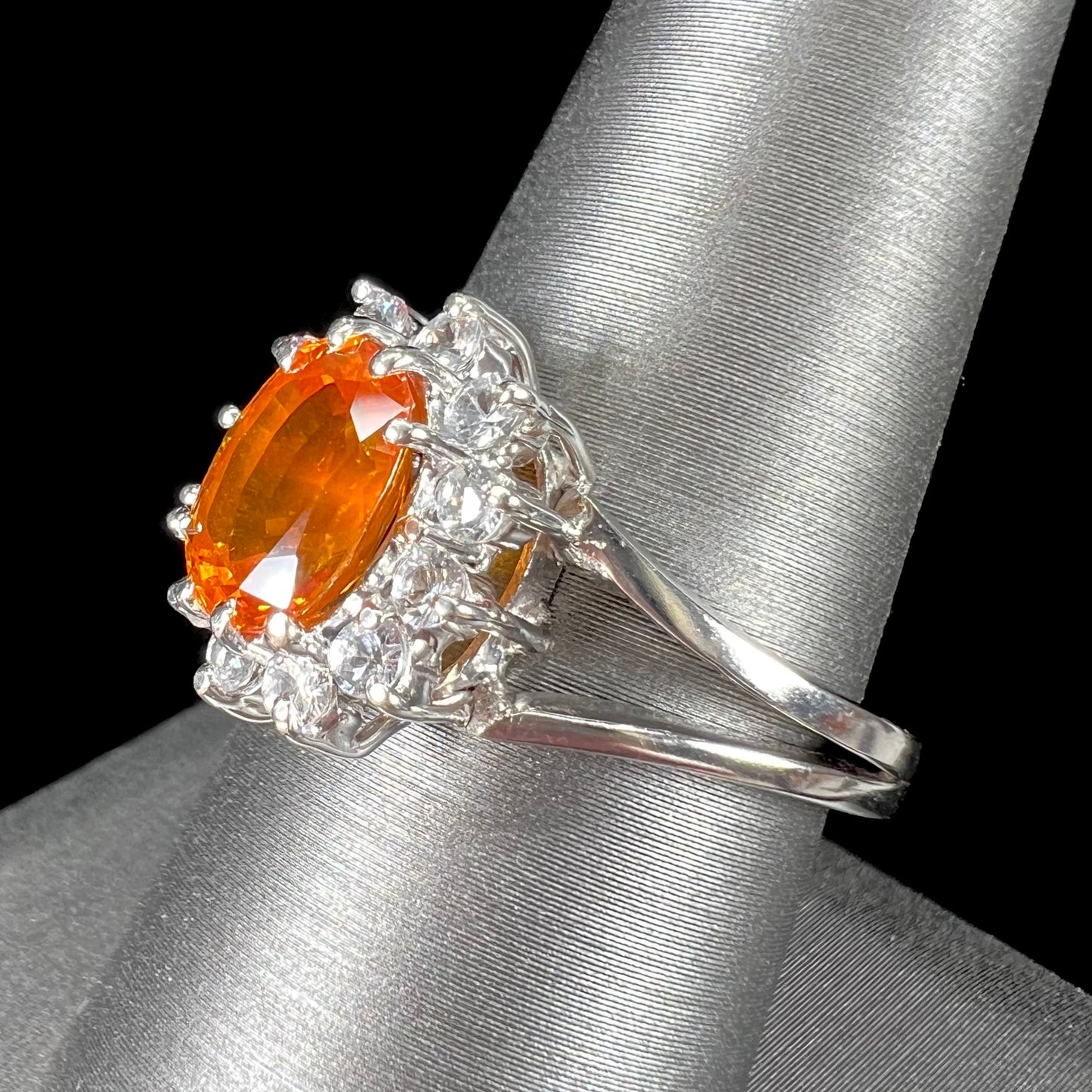 A white gold split shank ring set with an oval cut orange sapphire and a halo of white sapphires.