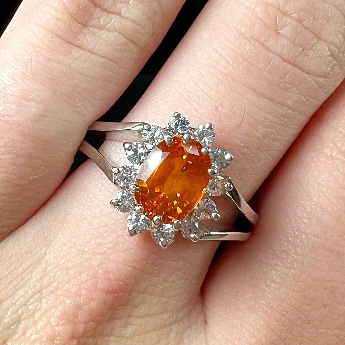 A white gold split shank ring set with an oval cut orange sapphire and a halo of white sapphires.