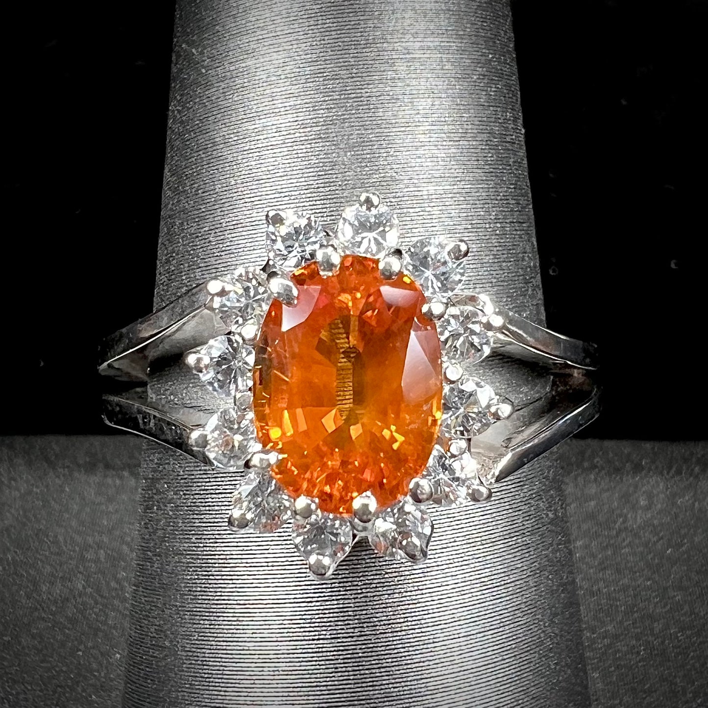 A white gold split shank ring set with an oval cut orange sapphire and a halo of white sapphires.