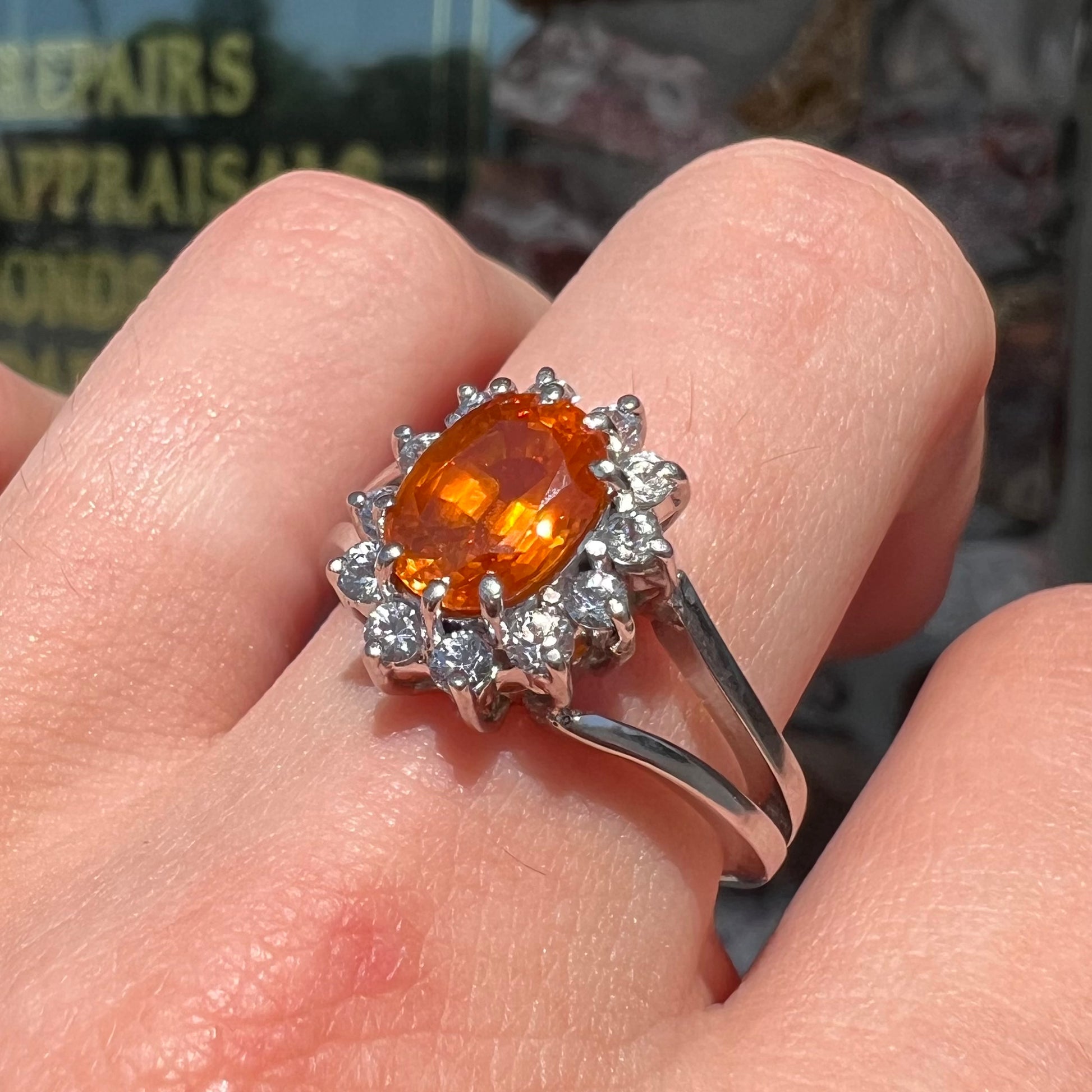 A white gold split shank ring set with an oval cut orange sapphire and a halo of white sapphires.