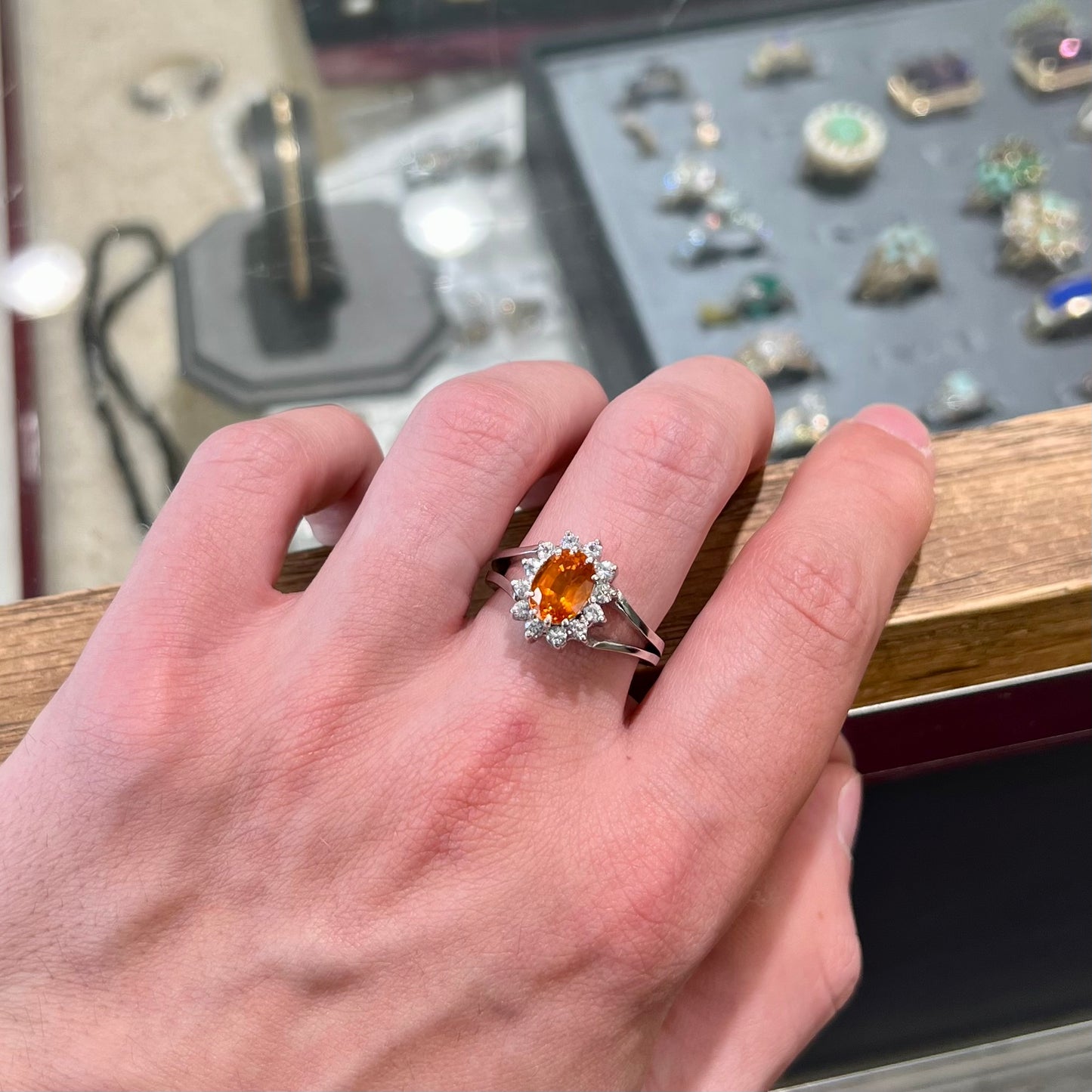 A white gold split shank ring set with an oval cut orange sapphire and a halo of white sapphires.