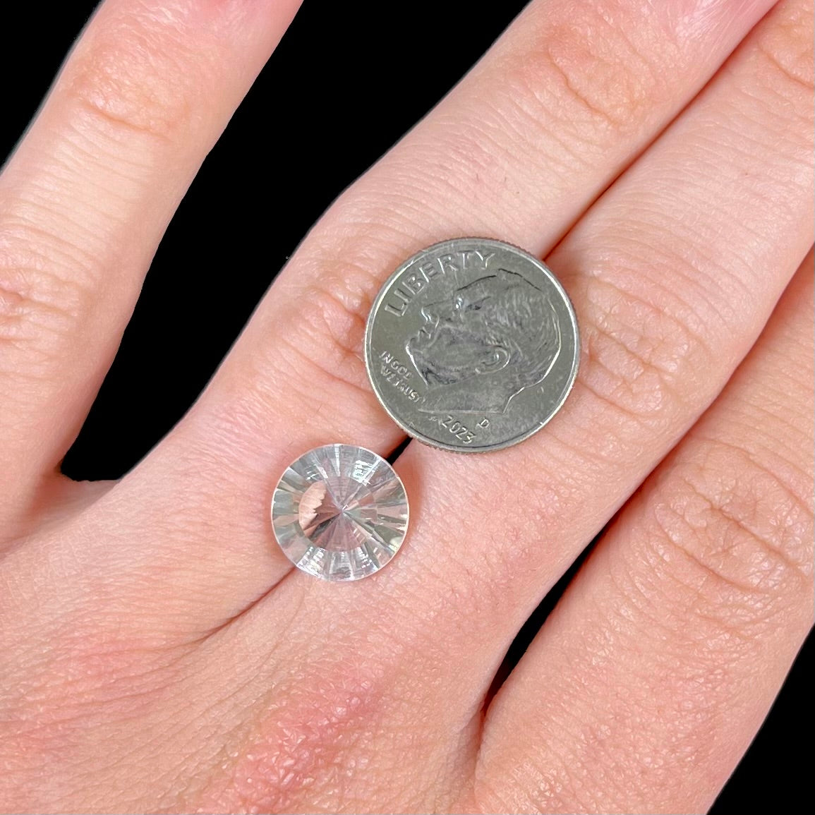 A loose, round cut petalite gemstone. The faceted stone is colorless with a black surface inclusion.