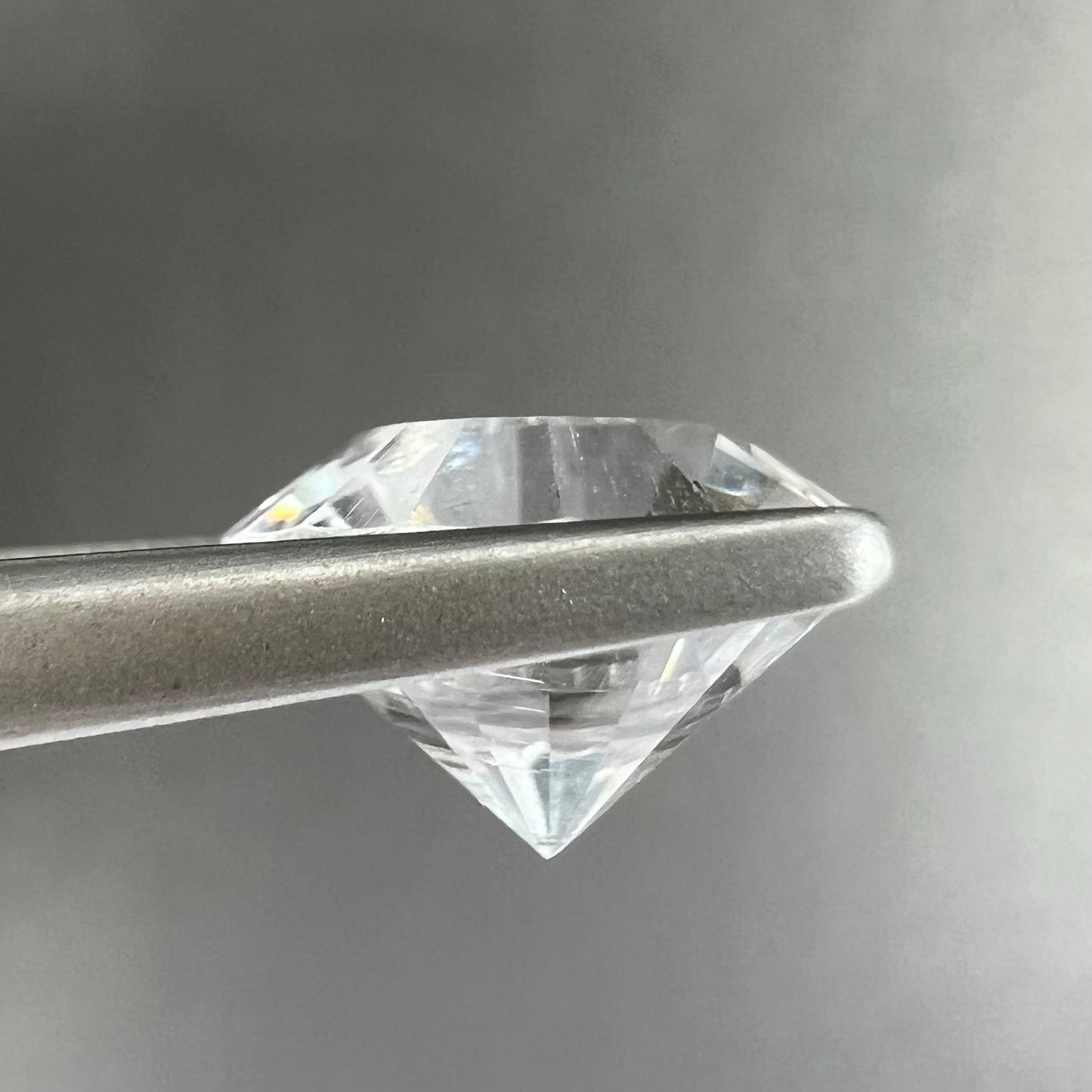 A loose, round cut petalite gemstone. The faceted stone is colorless with a black surface inclusion.