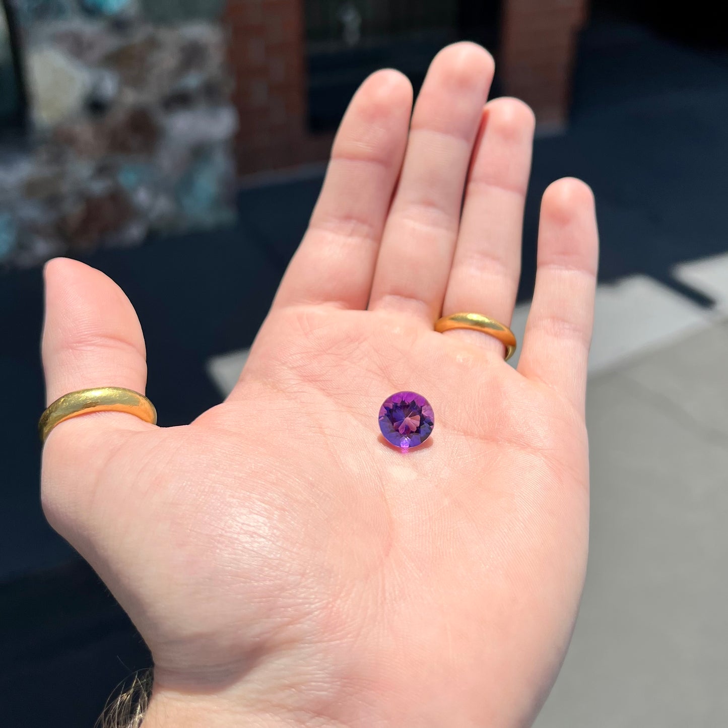 A loose, faceted round brilliant cut amethyst gemstone. The stone is purple with blue flashes.