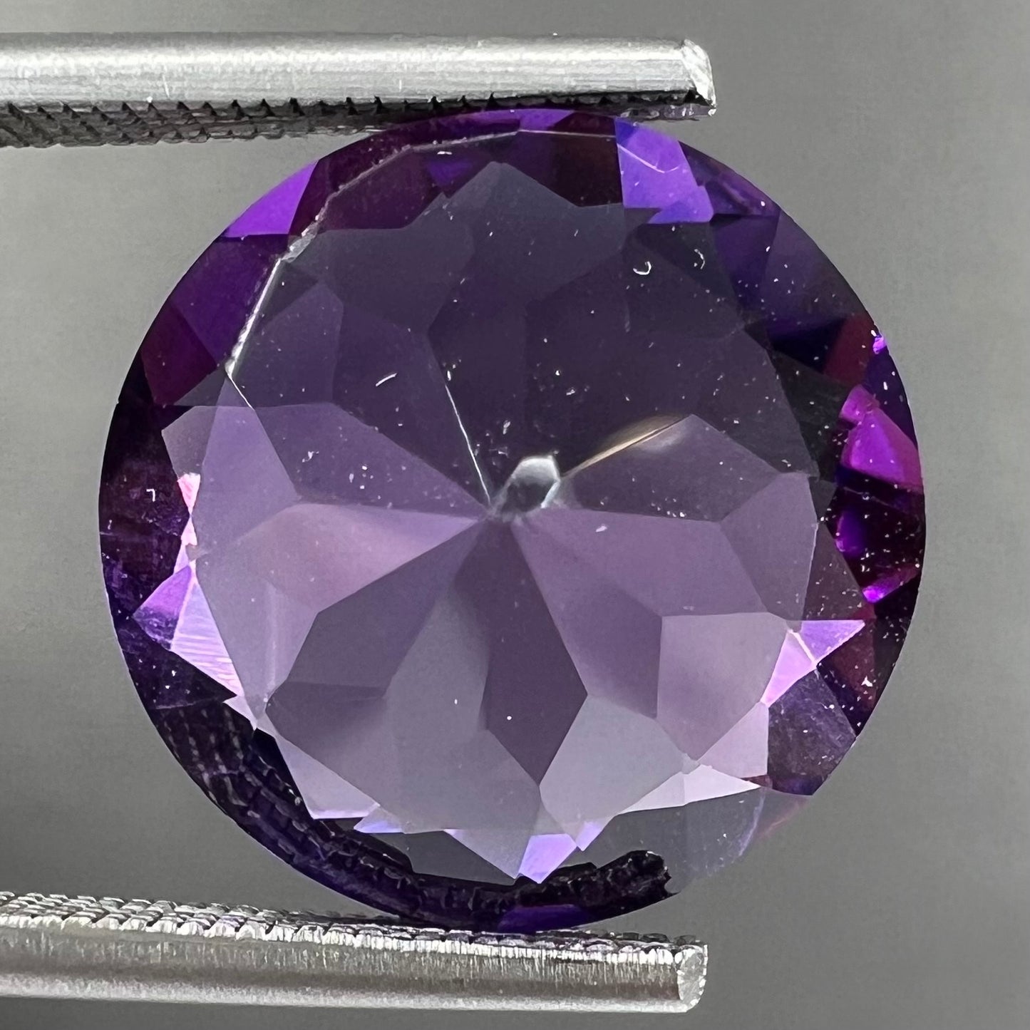 A loose, faceted round brilliant cut amethyst gemstone. The stone is purple with blue flashes.