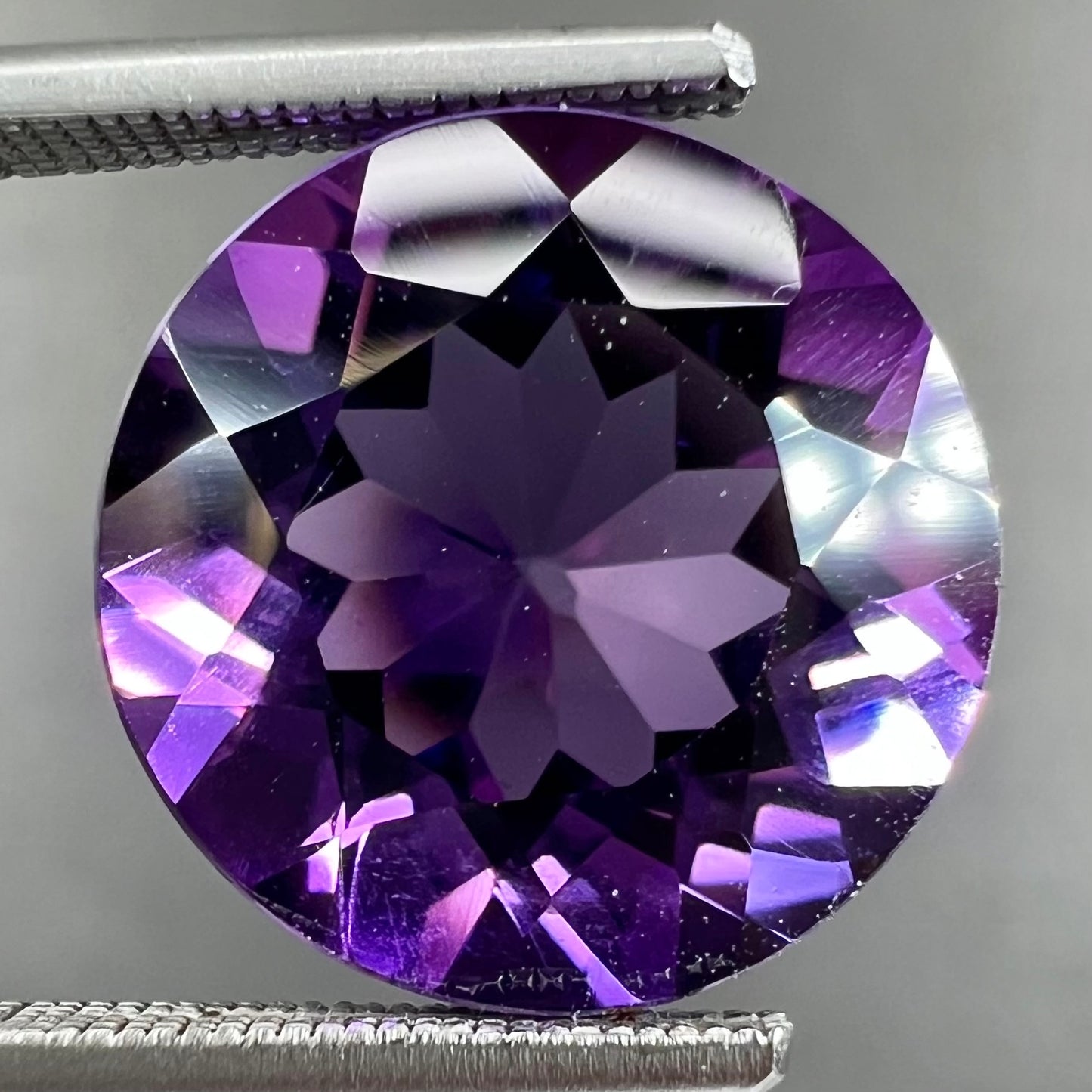 A loose, faceted round brilliant cut amethyst gemstone. The stone is purple with blue flashes.