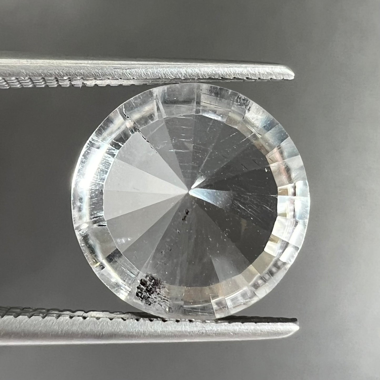 A loose, round cut petalite gemstone. The faceted stone is colorless with a black surface inclusion.