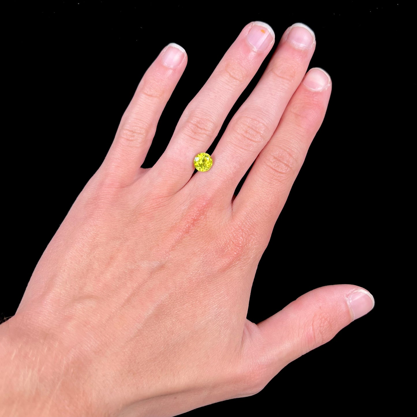 A loose, round brilliant cut sphene gemstone. The stone is green with orange, green, blue, and yellow flashes.