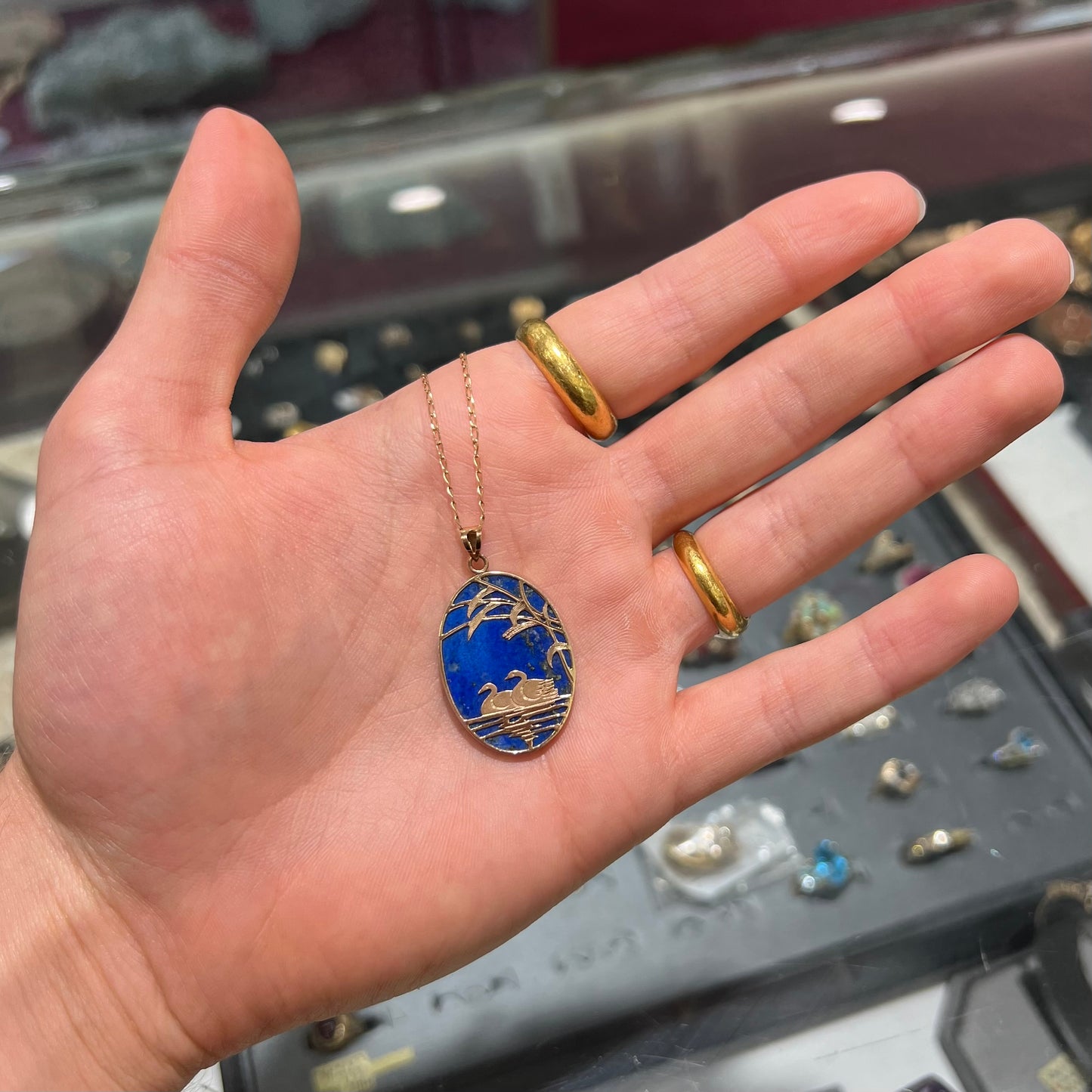 An oval cut lapis lazuli pendant set in a yellow gold frame featuring the design of two swans swimming in a pond.