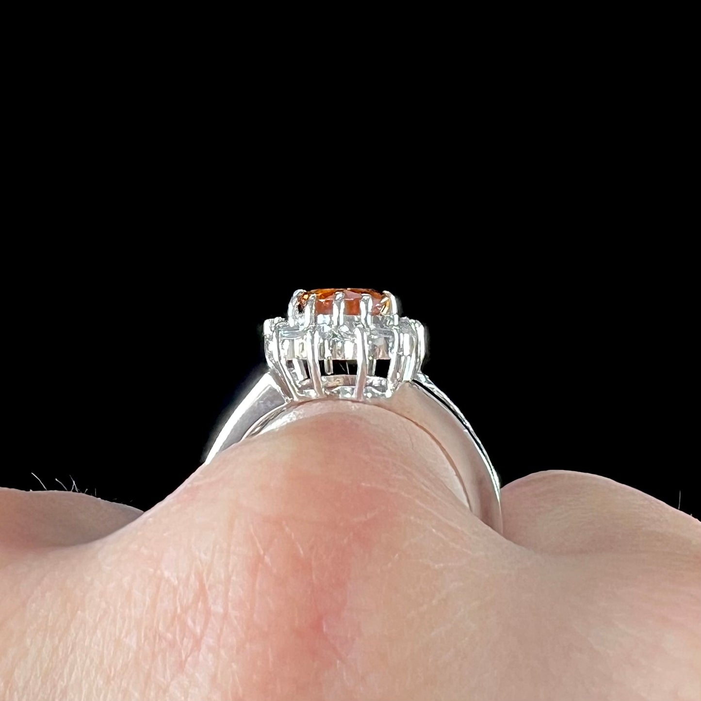 A white gold split shank ring set with an oval cut orange sapphire and a halo of white sapphires.