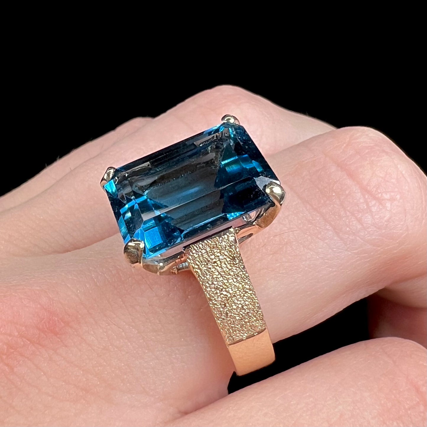 A ladies' yellow gold London blue topaz solitaire ring. The ring is textured, and the topaz is emerald cut.