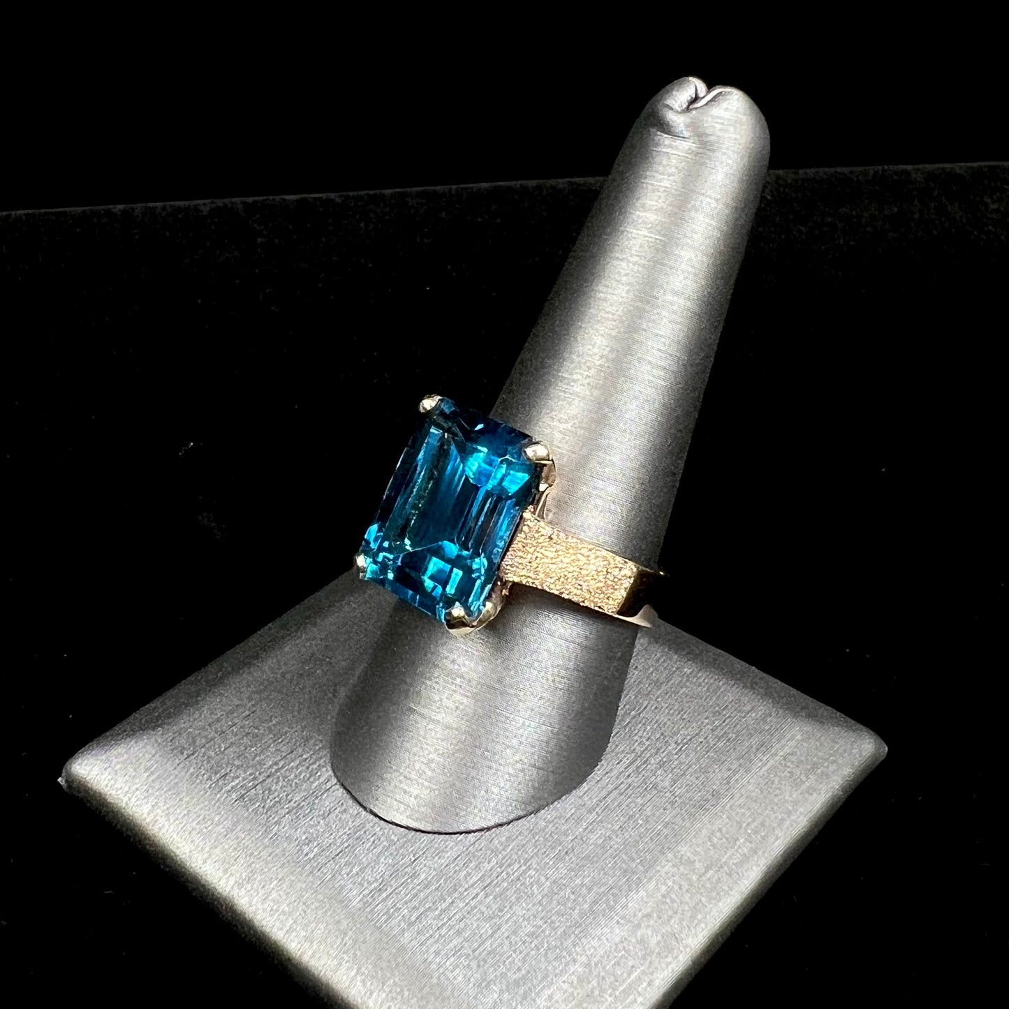 A ladies' yellow gold London blue topaz solitaire ring. The ring is textured, and the topaz is emerald cut.