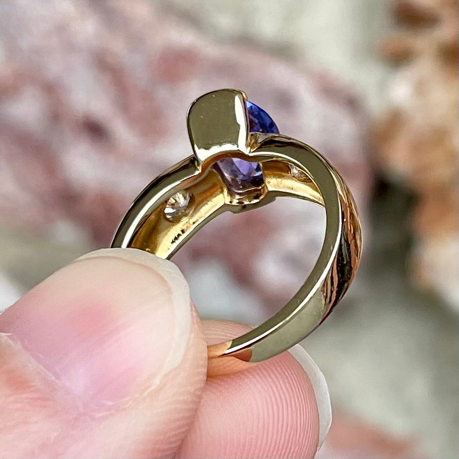 A yellow gold ring half-bezel set with an oval cut blue tanzanite and two flush set round diamonds.