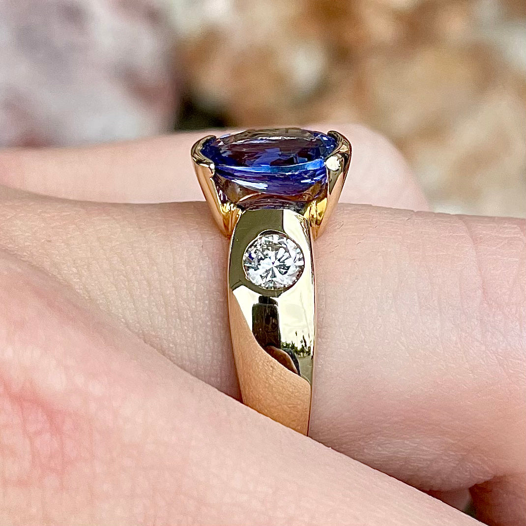 A yellow gold ring half-bezel set with an oval cut blue tanzanite and two flush set round diamonds.