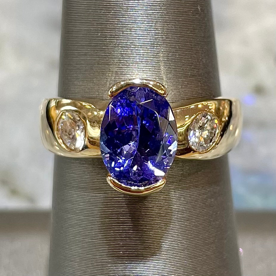 A yellow gold ring half-bezel set with an oval cut blue tanzanite and two flush set round diamonds.