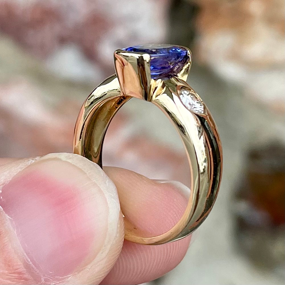 A yellow gold ring half-bezel set with an oval cut blue tanzanite and two flush set round diamonds.