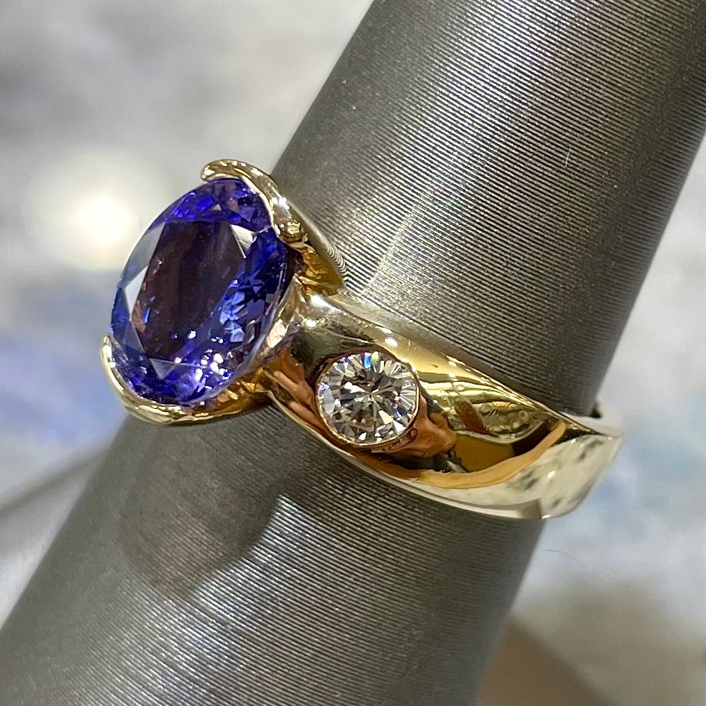 A yellow gold ring half-bezel set with an oval cut blue tanzanite and two flush set round diamonds.